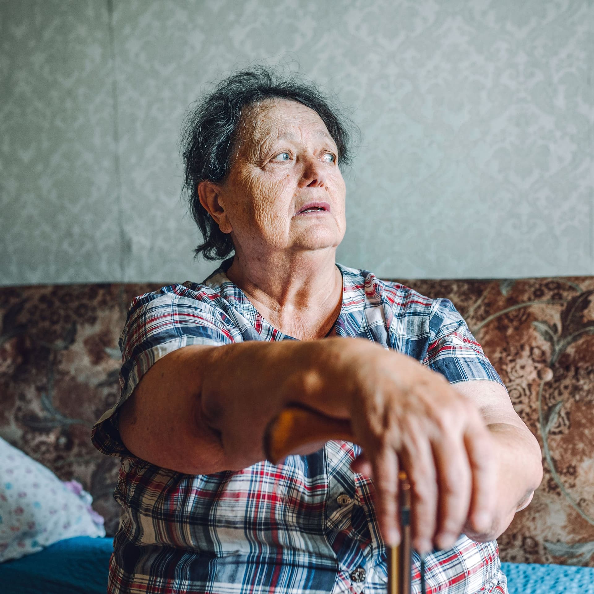 Eine ältere Frau sitzt auf einem Sofa und stützt sich mit den Händen auf einen Gehstock.