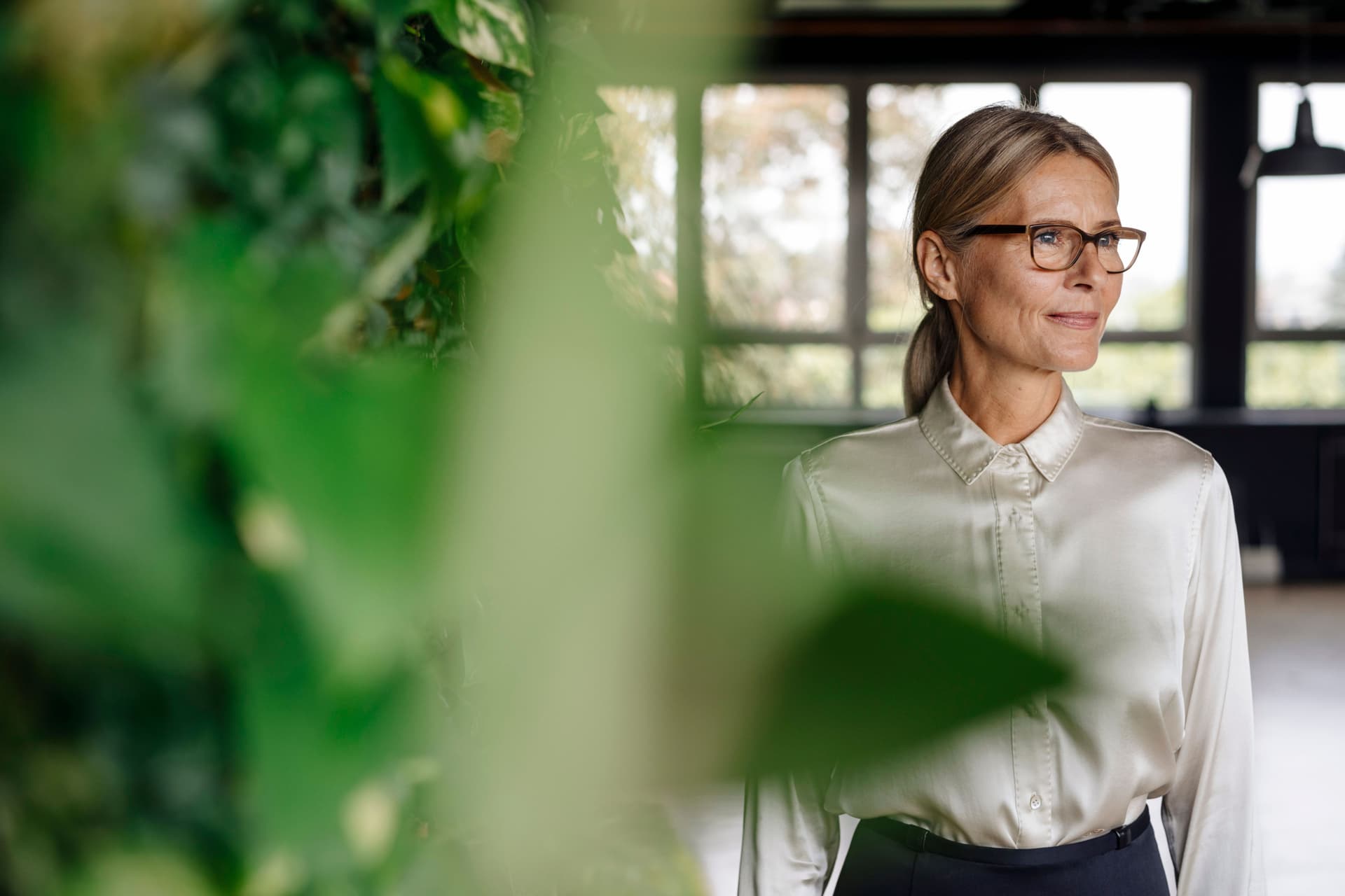 Lächelnde mittelalte Frau mit Brille und schicker Bluse im Büro guckt zur Seite. Im Vordergrund sind grüne Pflanzen.