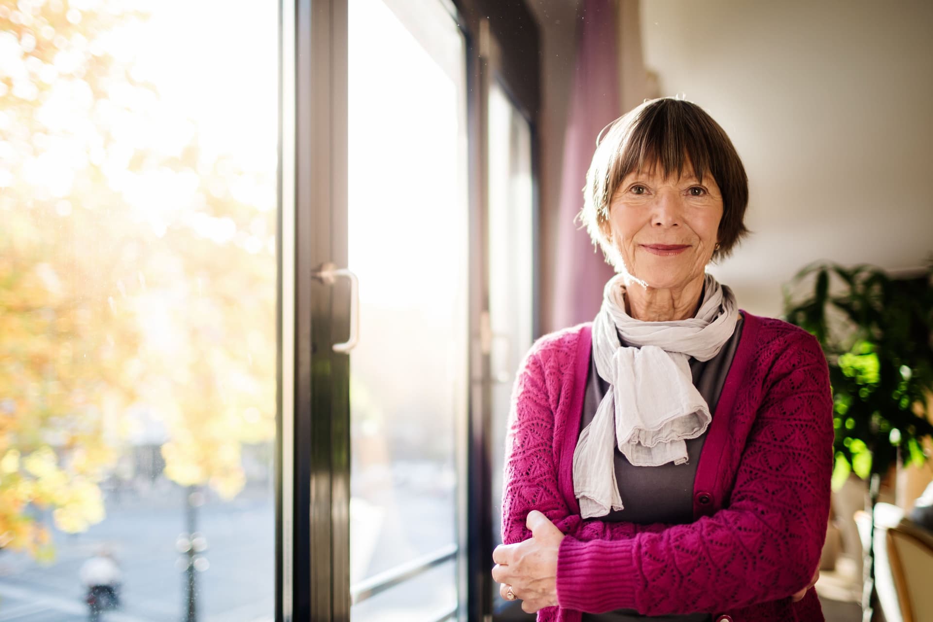Ältere Frau, die am Fenster steht und in die Kamera lächelt. Der Blick durch das Fenster zeigt Bäume, die in Herbstfarben in der Sonne leuchten.
