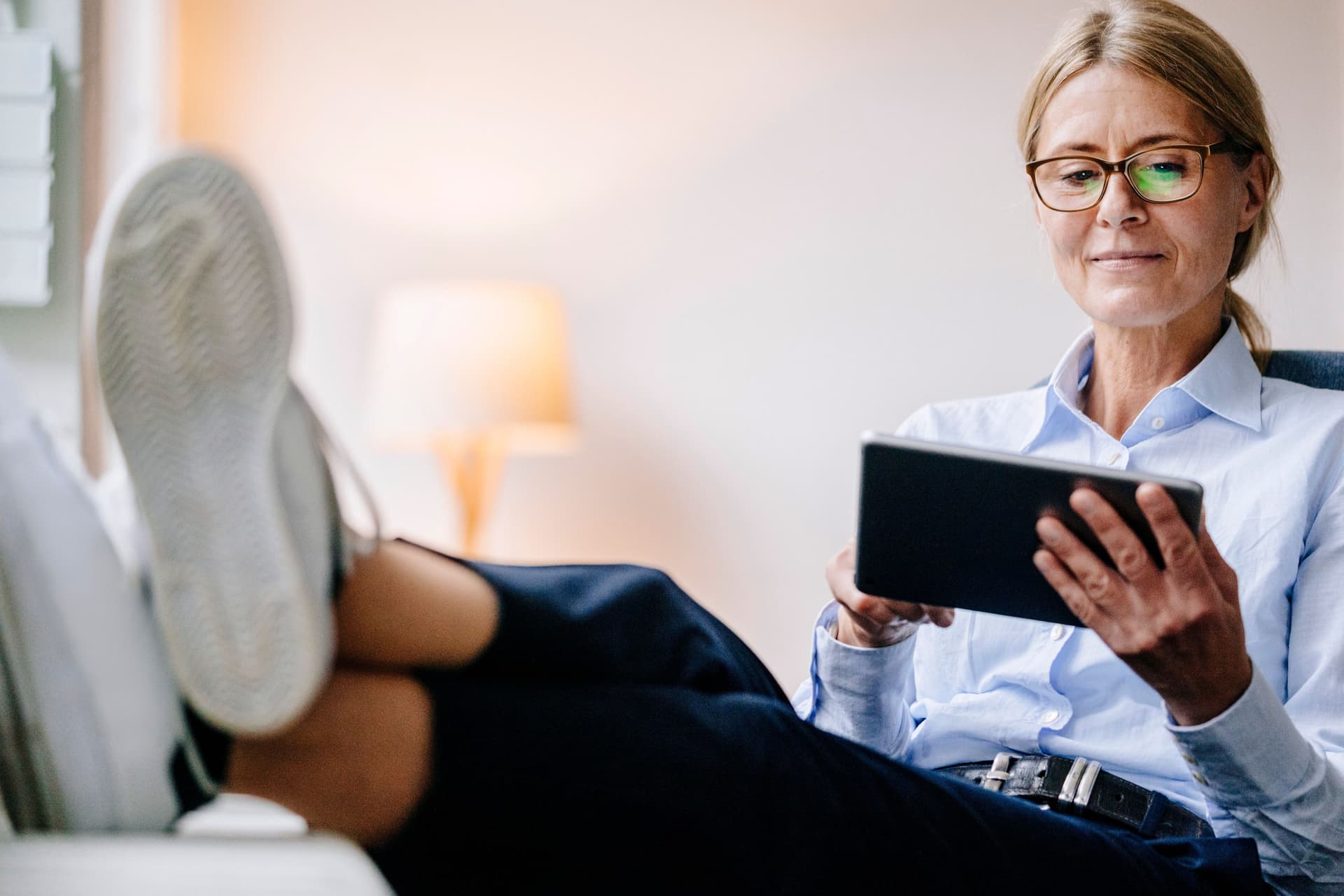Eine Frau mittleren Alters hält ein Tablet in der Hand und legt entspannt ihre Füße auf den Tisch.