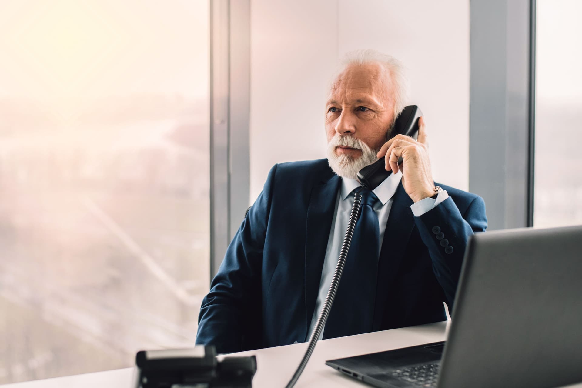 Älterer Mann in Anzug am Telefon mit einem Laptop vor sich in einem Büro. Hinter ihm ist eine Fensterfront mit einer Skyline.