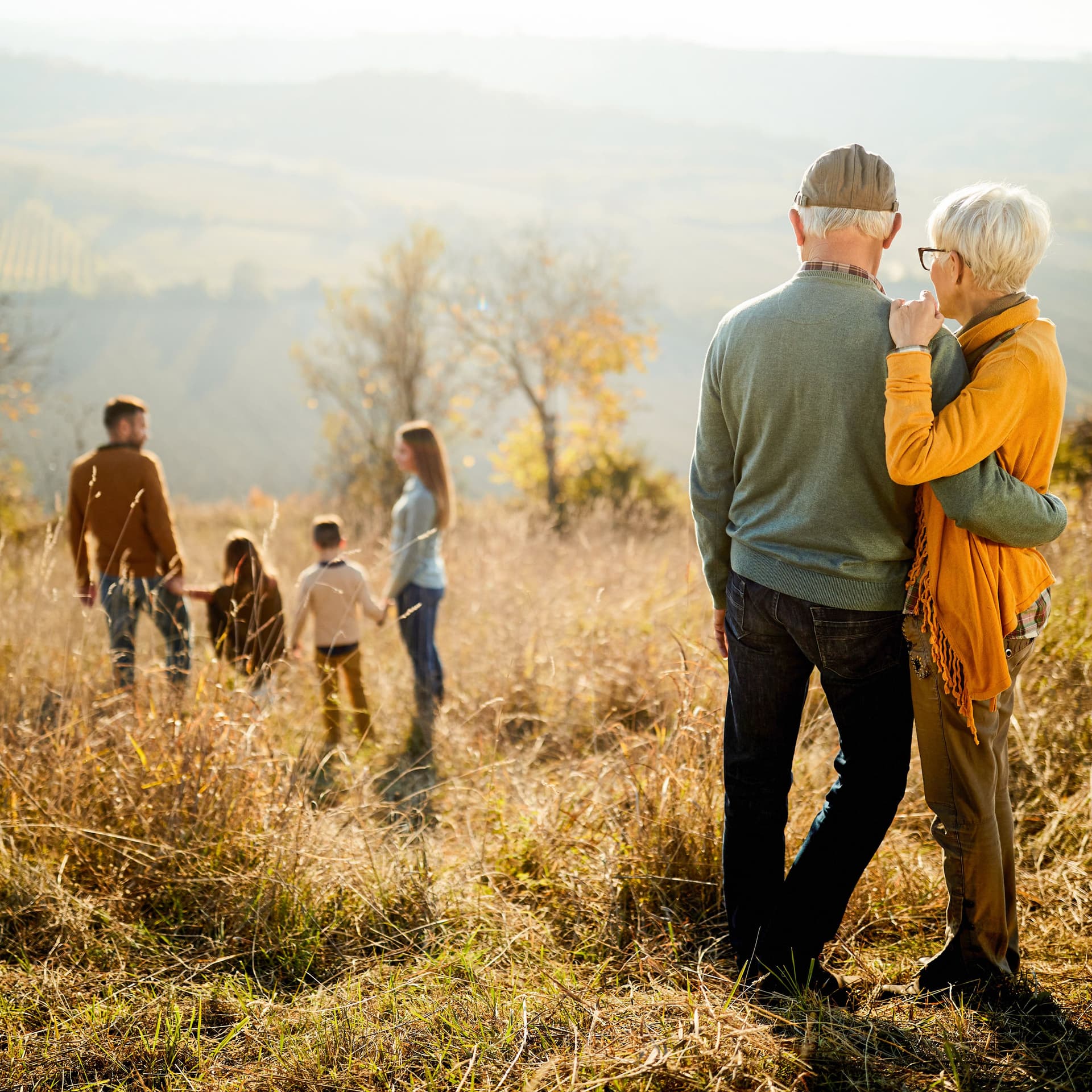 Älteres Paar steht Arm in Arm auf einer hoch gelegenen Wiese und blickt auf eine jüngere Familie im Hintergrund.