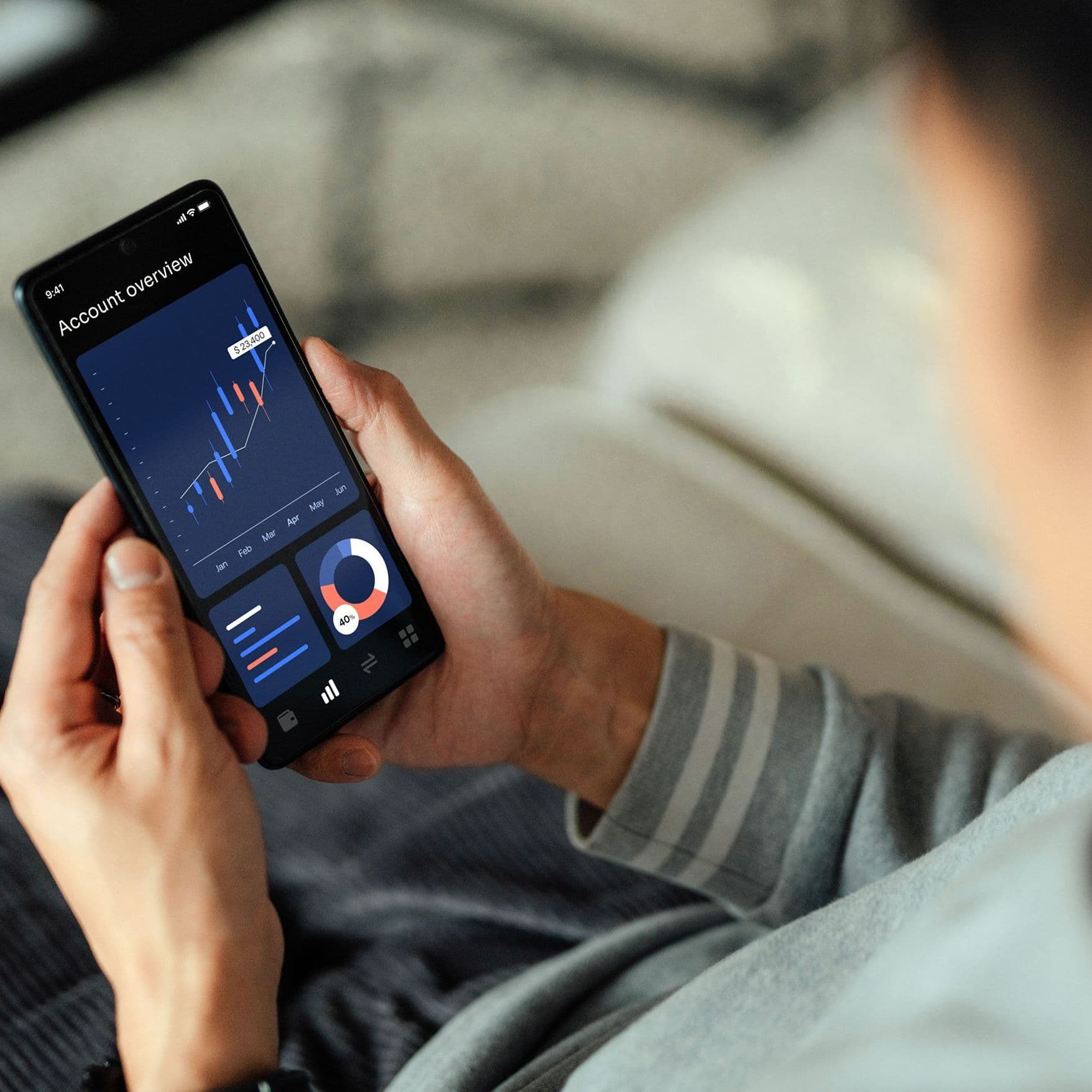 Nahaufnahme eines Smartphones, welches ein Mann zuhause auf dem Sofa sitzend, hält. Der Screen zeigt Aktienauswertungen, Aktienkurse, Aktienwerte in Form von Diagrammen. 