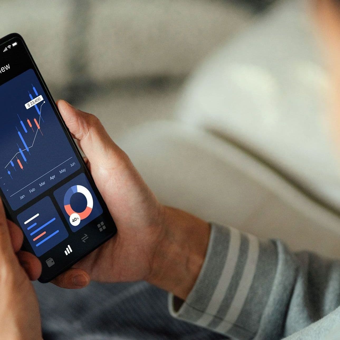 Nahaufnahme eines Smartphones, welches ein Mann zuhause auf dem Sofa sitzend, hält. Der Screen zeigt Aktienauswertungen, Aktienkurse, Aktienwerte in Form von Diagrammen. 