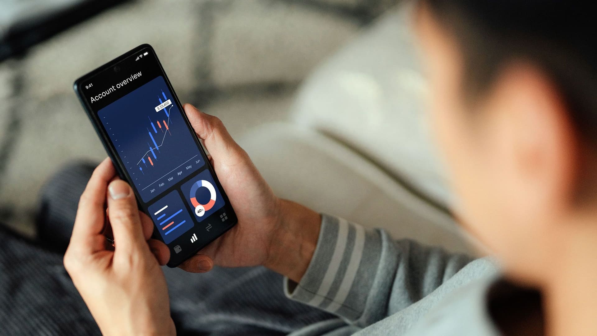 Nahaufnahme eines Smartphones, welches ein Mann zuhause auf dem Sofa sitzend, hält. Der Screen zeigt Aktienauswertungen, Aktienkurse, Aktienwerte in Form von Diagrammen. 