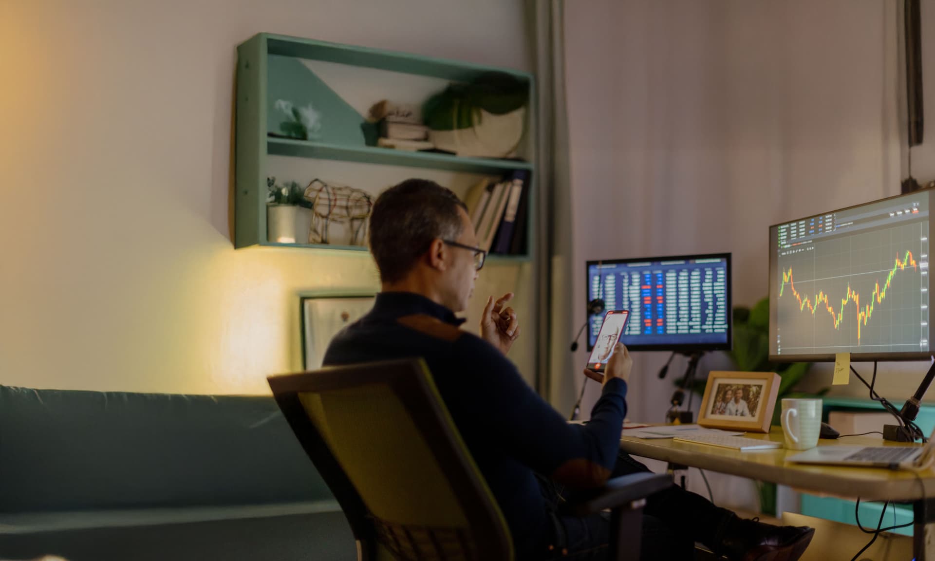EIn Mann sitzt am Schreibtisch und schaut auf sein Smartphone. Auf seinen Monitoren sind Handelskurse.