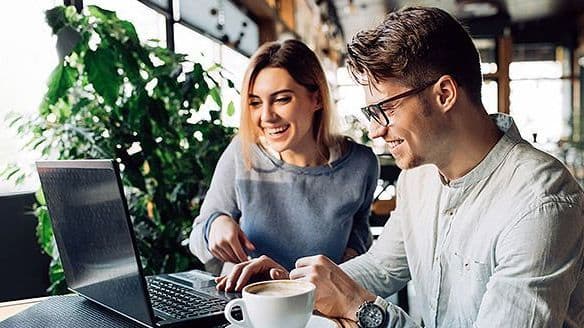 zwei junge Menschen schauen gemeinsam lächelnd auf einen Laptop in einem Café