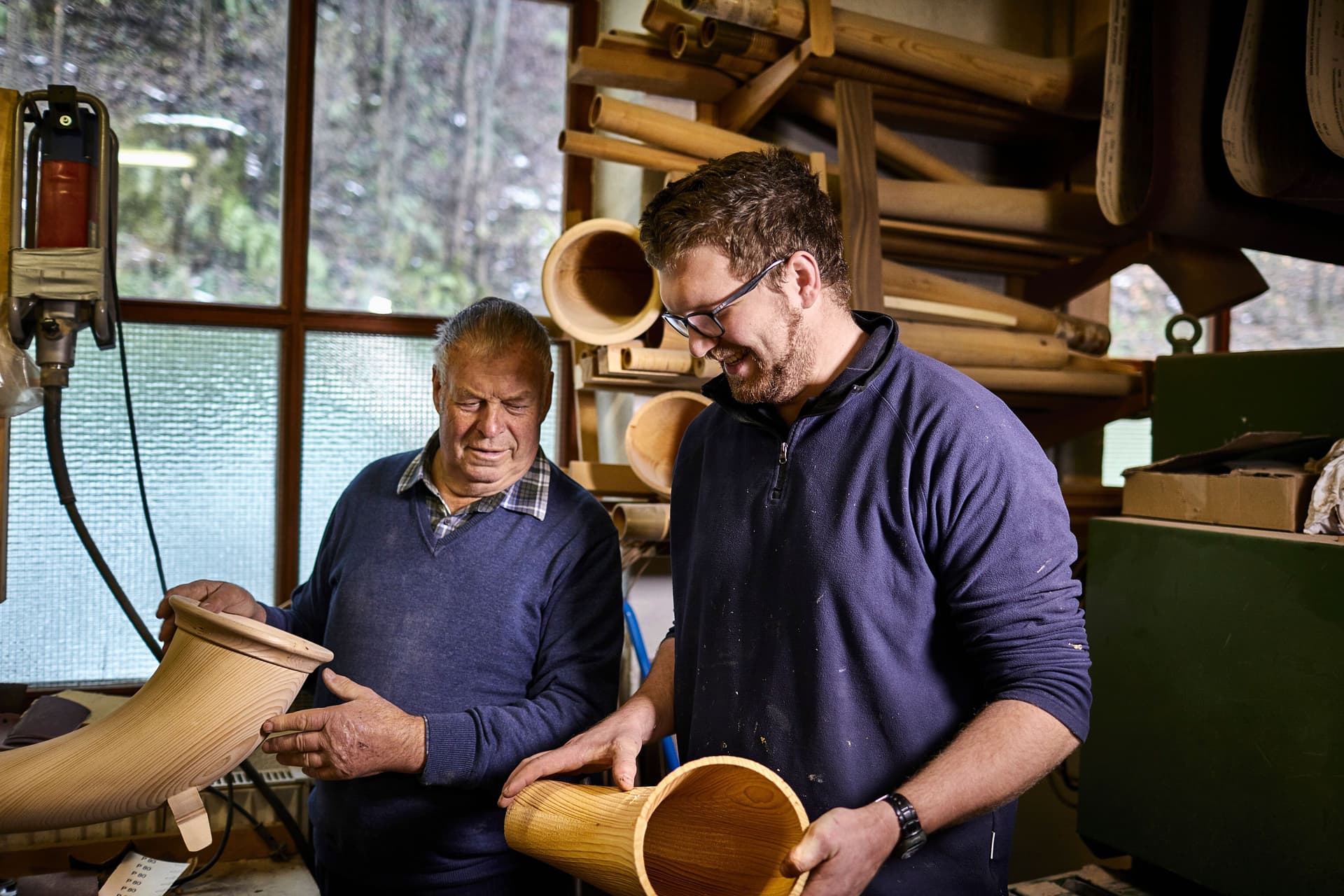 Alter und junger Instrumentenbauer in einer Werkstatt arbeiten an Alphörnern und lächeln dabei.