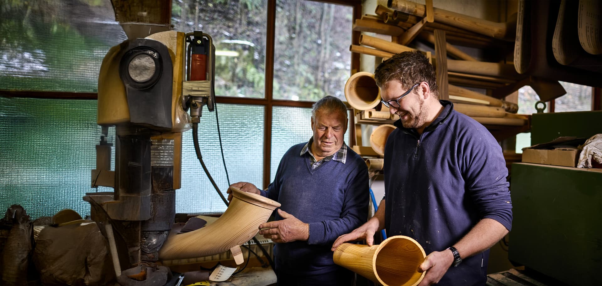 Alter und junger Instrumentenbauer in einer Werkstatt arbeiten an Alphörnern und lächeln dabei.