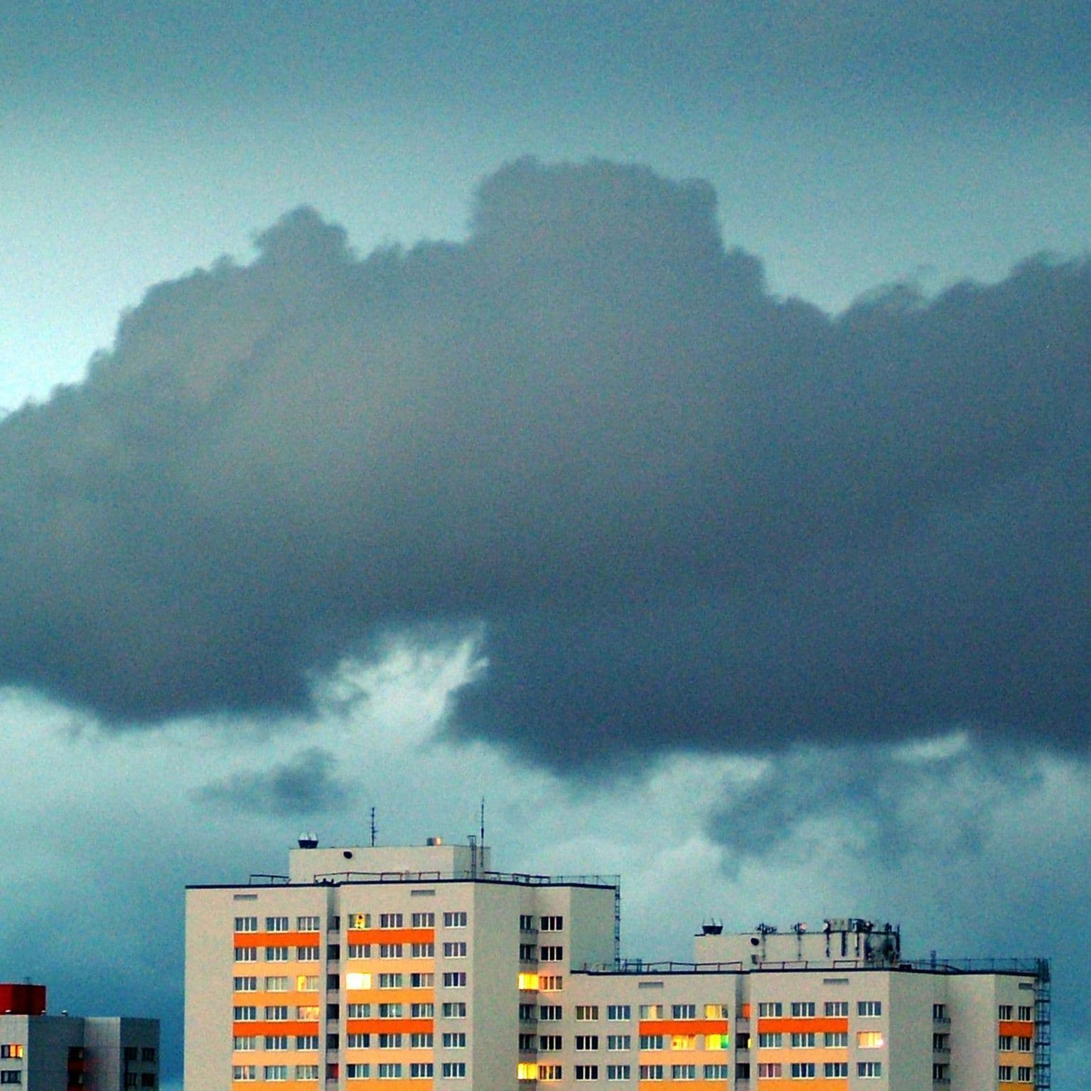 Dunkle Wolken in einem weiten Himmel ziehen über Hochhäuser deren Fenster beleuchtet sind.