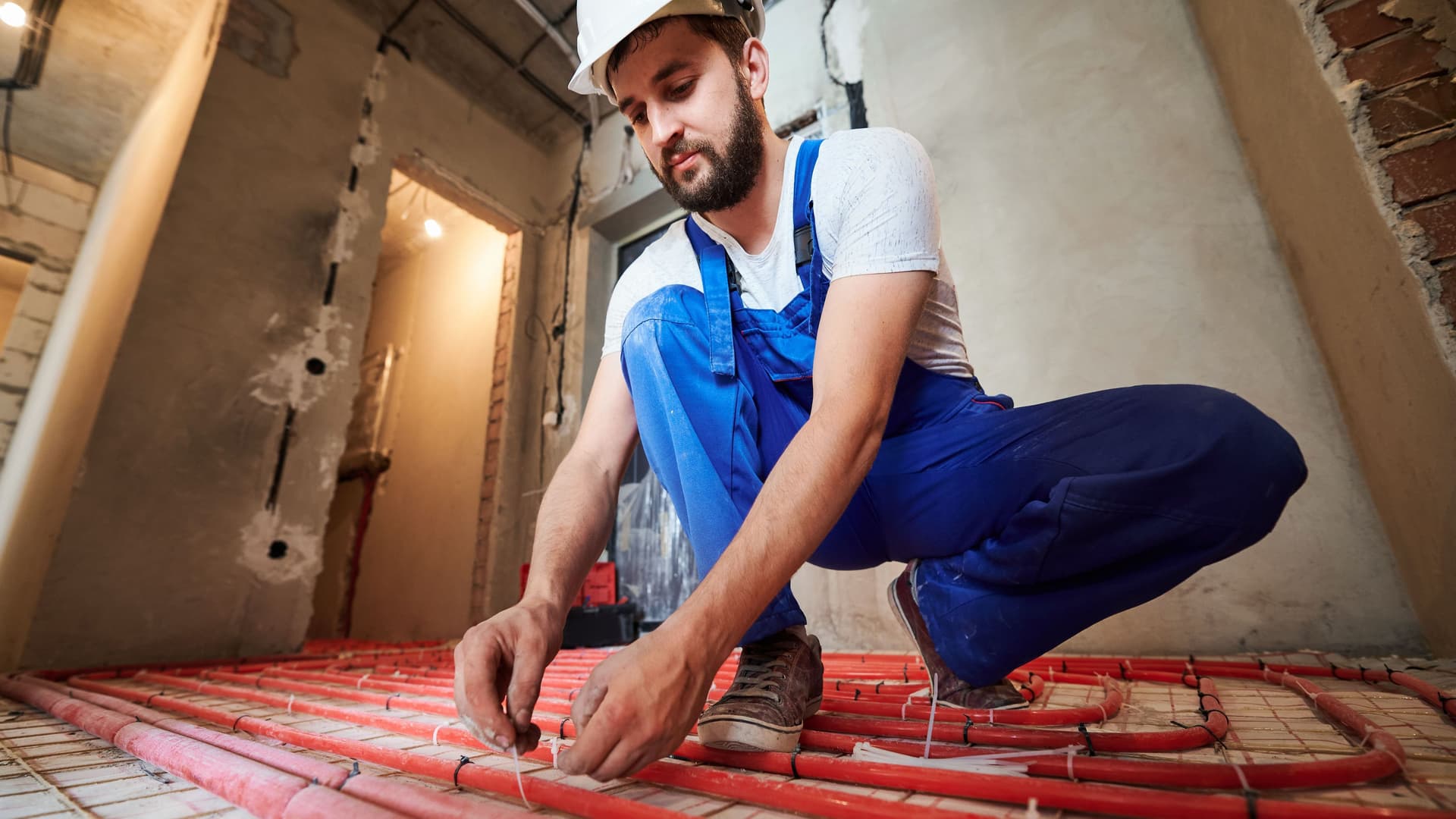 Mann mit blauer Arbeitslatzhose und weißem Schutzhelm installiert eine Fußbodenheizung.