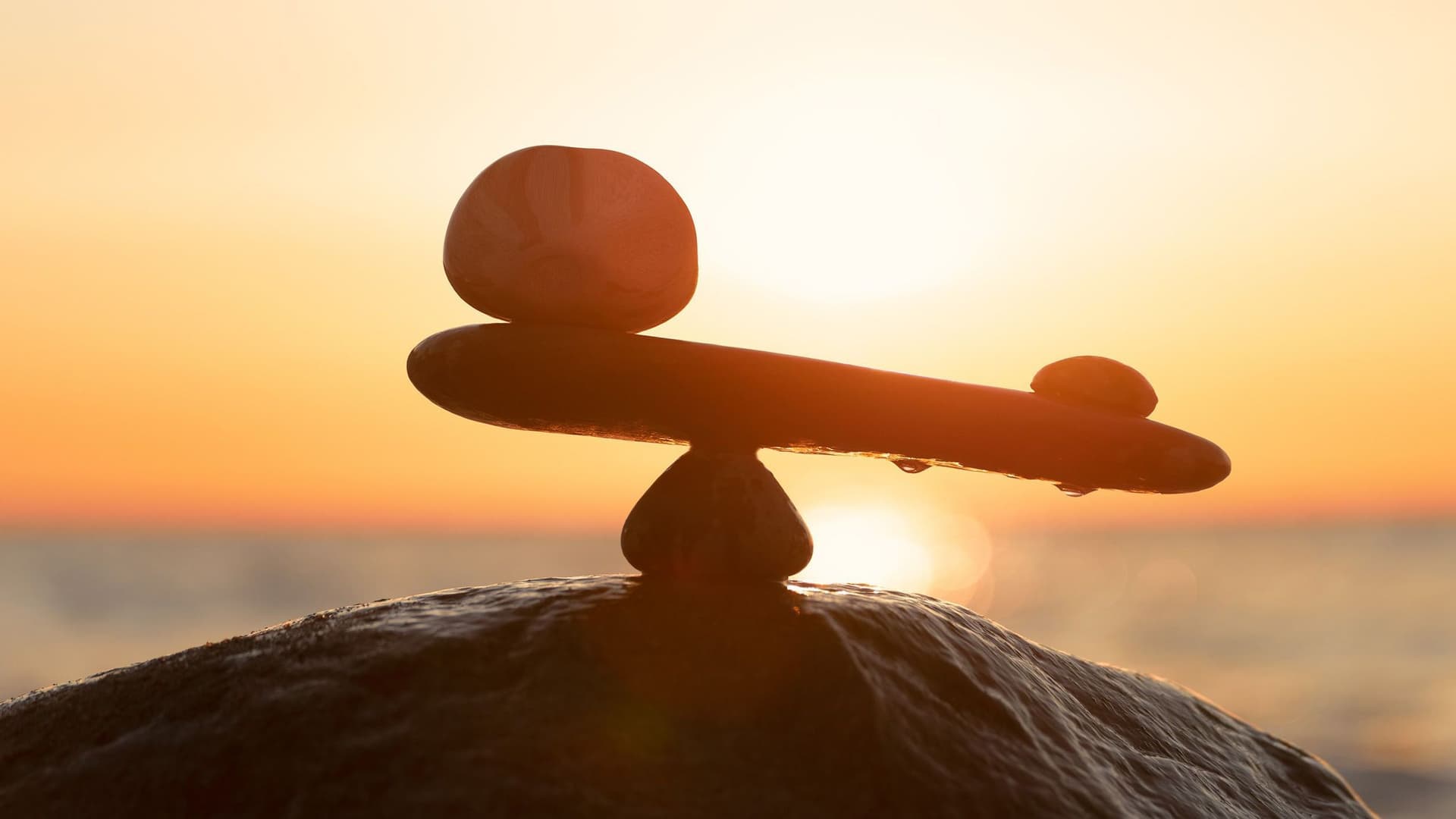 Auf dem Bild sind mehrere Steine ​​zu sehen, die in einer Balance angeordnet sind. Im Hintergrund geht die Sonne unter, und das warme Licht erzeugt eine ruhige, natürliche Stimmung.