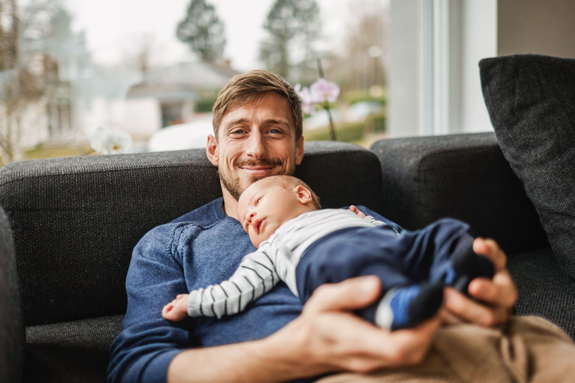 Ein lächelnder Vater liegt mit seinem Baby auf der Couch.