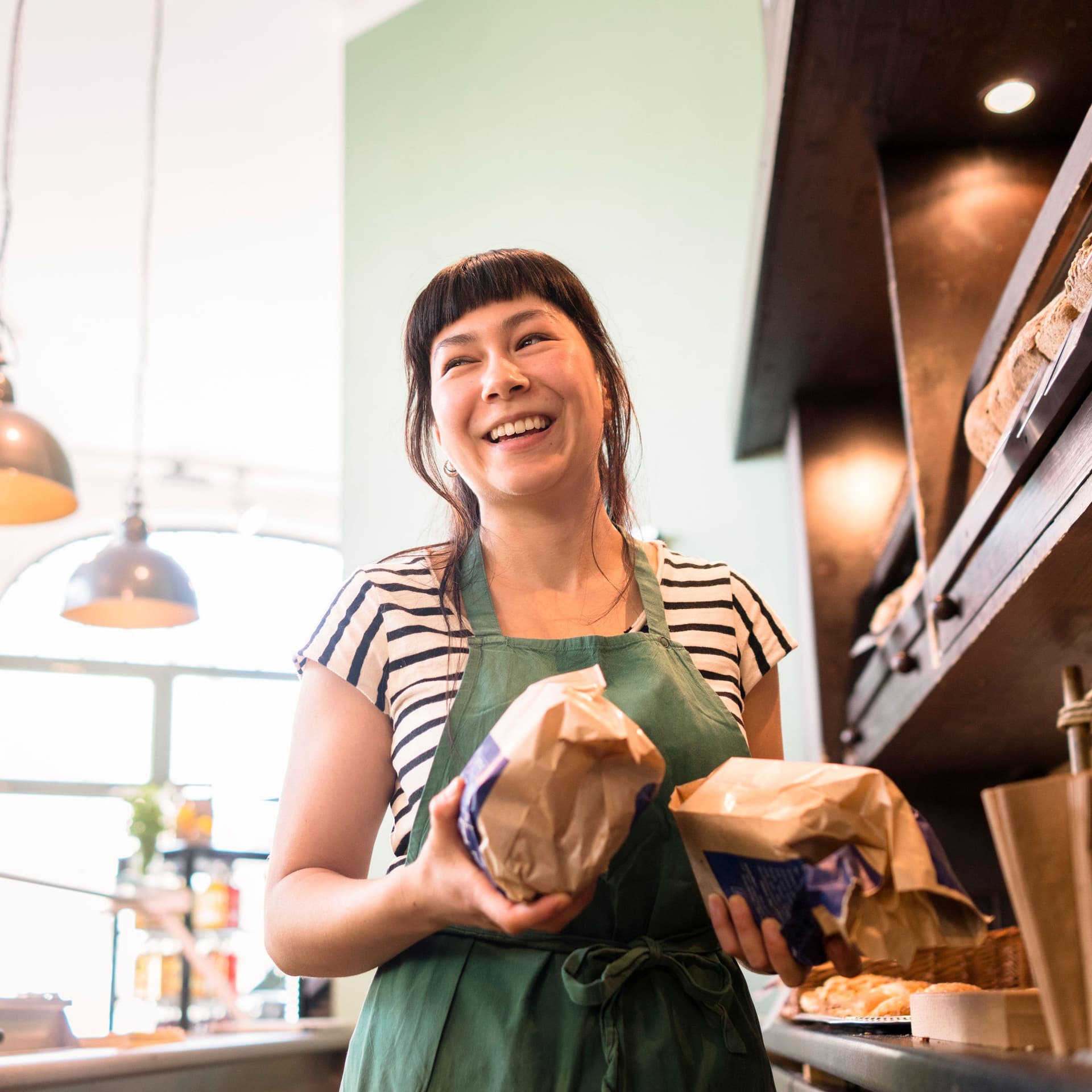 Lächelnde Besitzerin mit zwei eingepackten Broten in der Hand steht  zwischen einer Theke und Holzregalen in einer Bäckerei.