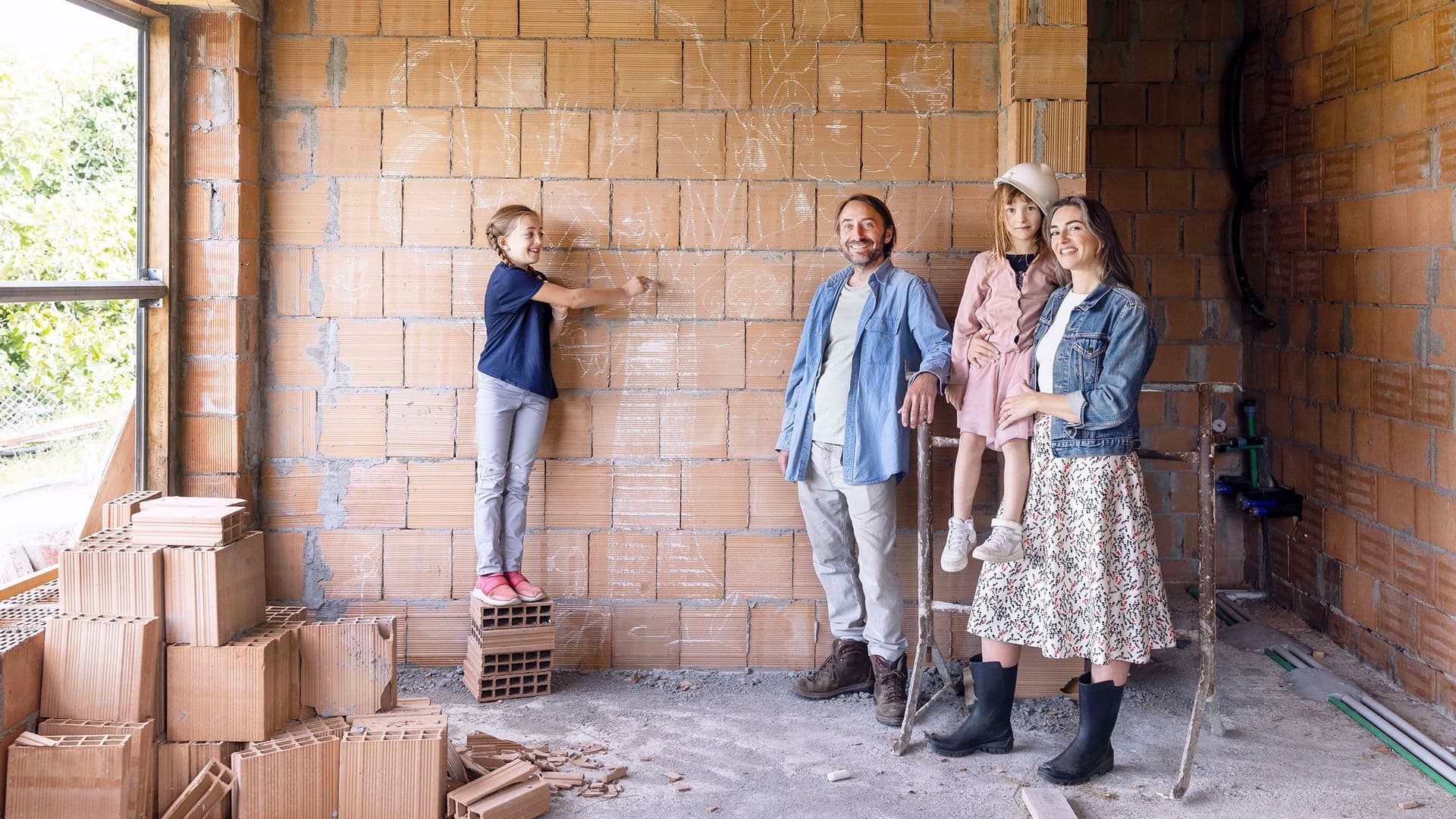 Eine vierköpfige Familie steht in dem Rohbau ihres neuen Hauses vor einer unverputzten Mauersteinwand und schaut in die Kamera. 