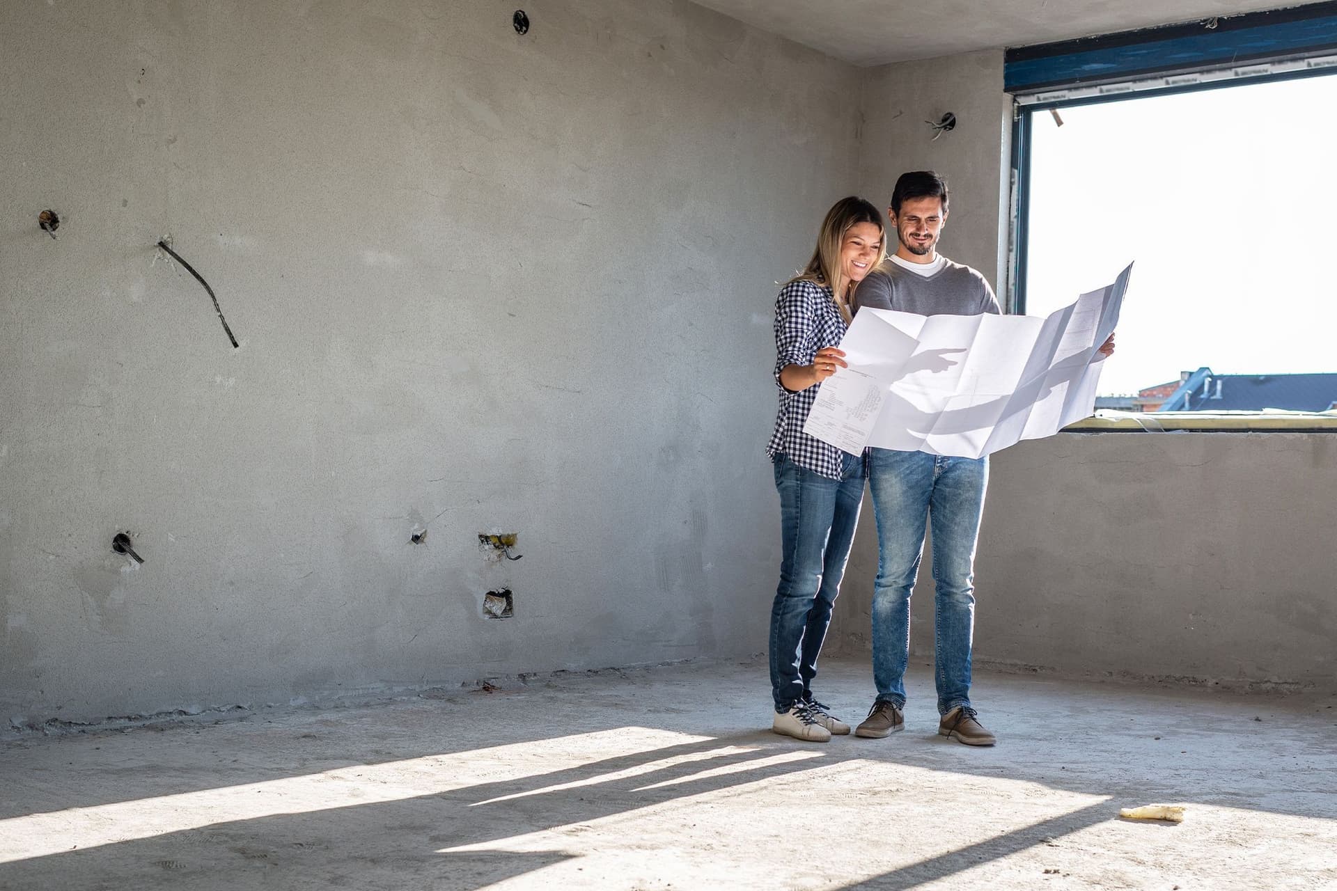 Eine Frau und ein Mann stehen inmitten eines Rohbaus auf einer Baustelle und sehen sich zusammen den Bauplan an. 