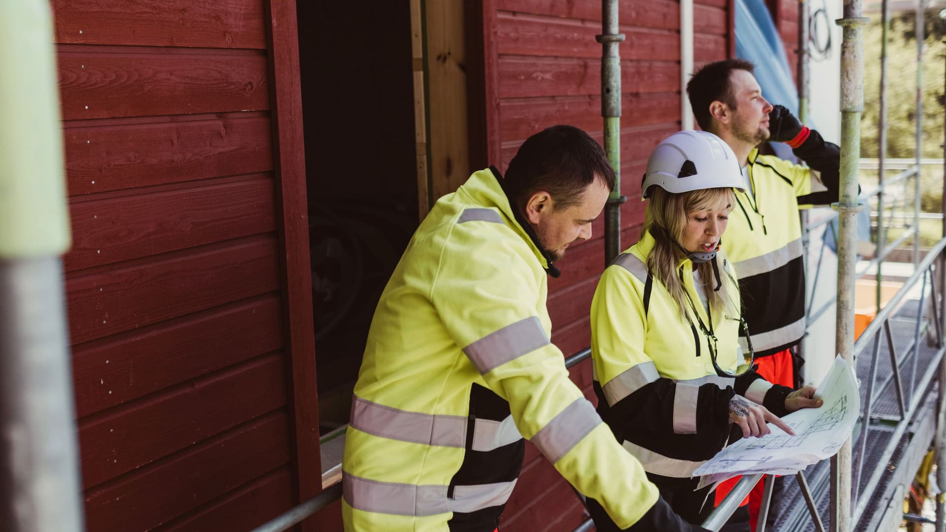 Eine Frau und ein Mann in gelber Arbeitsbekleidung besprechen einen Bauplan. Im Hintergrund ist ein weiterer Mann in gelber Arbeitskleidung telefonierend zu erkennen.