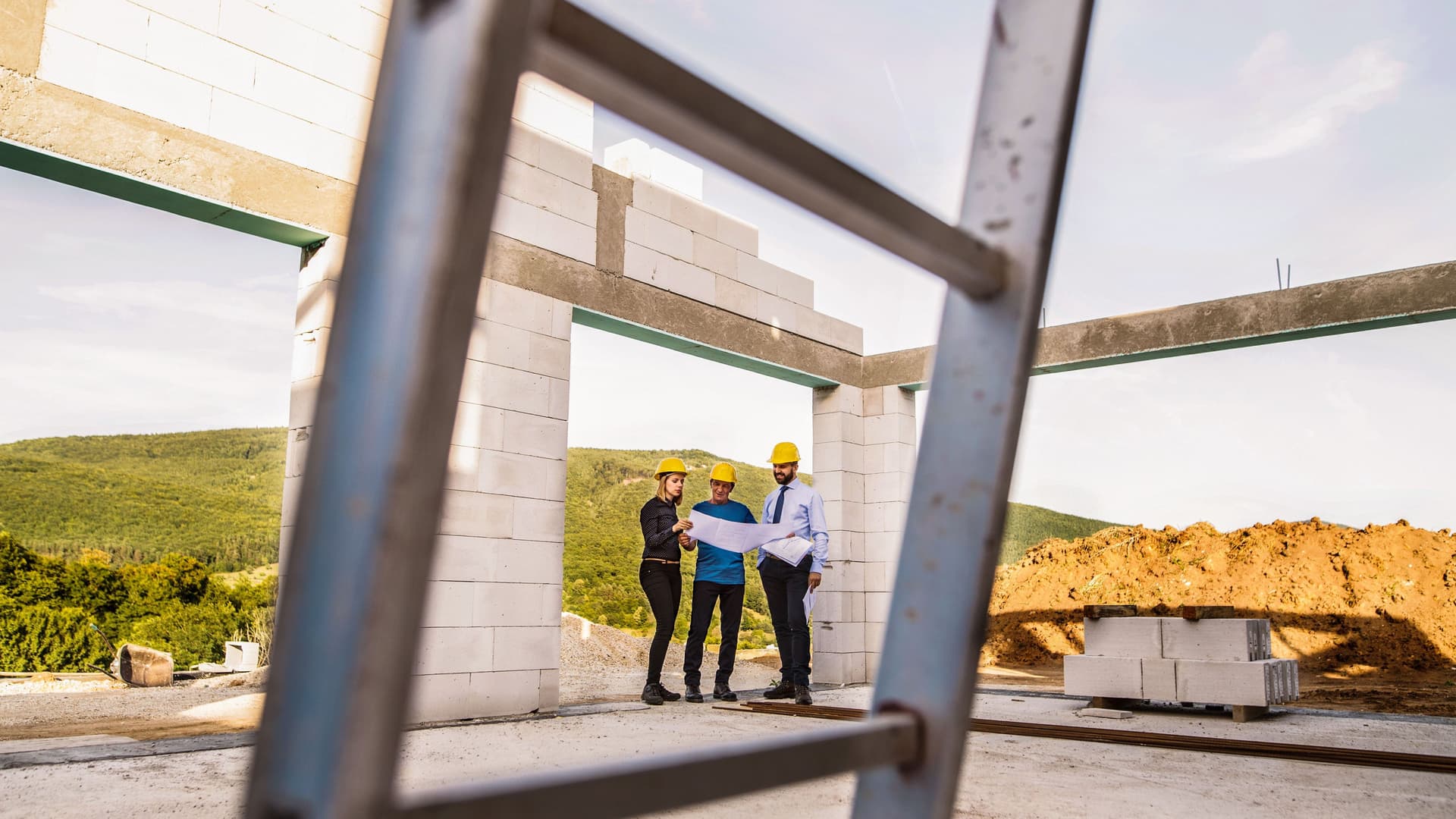 Drei Personen stehen mit gelben Helmen und Plänen auf Papier auf einer Baustelle eines Hauses. Im Vordergrund steht eine Metallleiter.