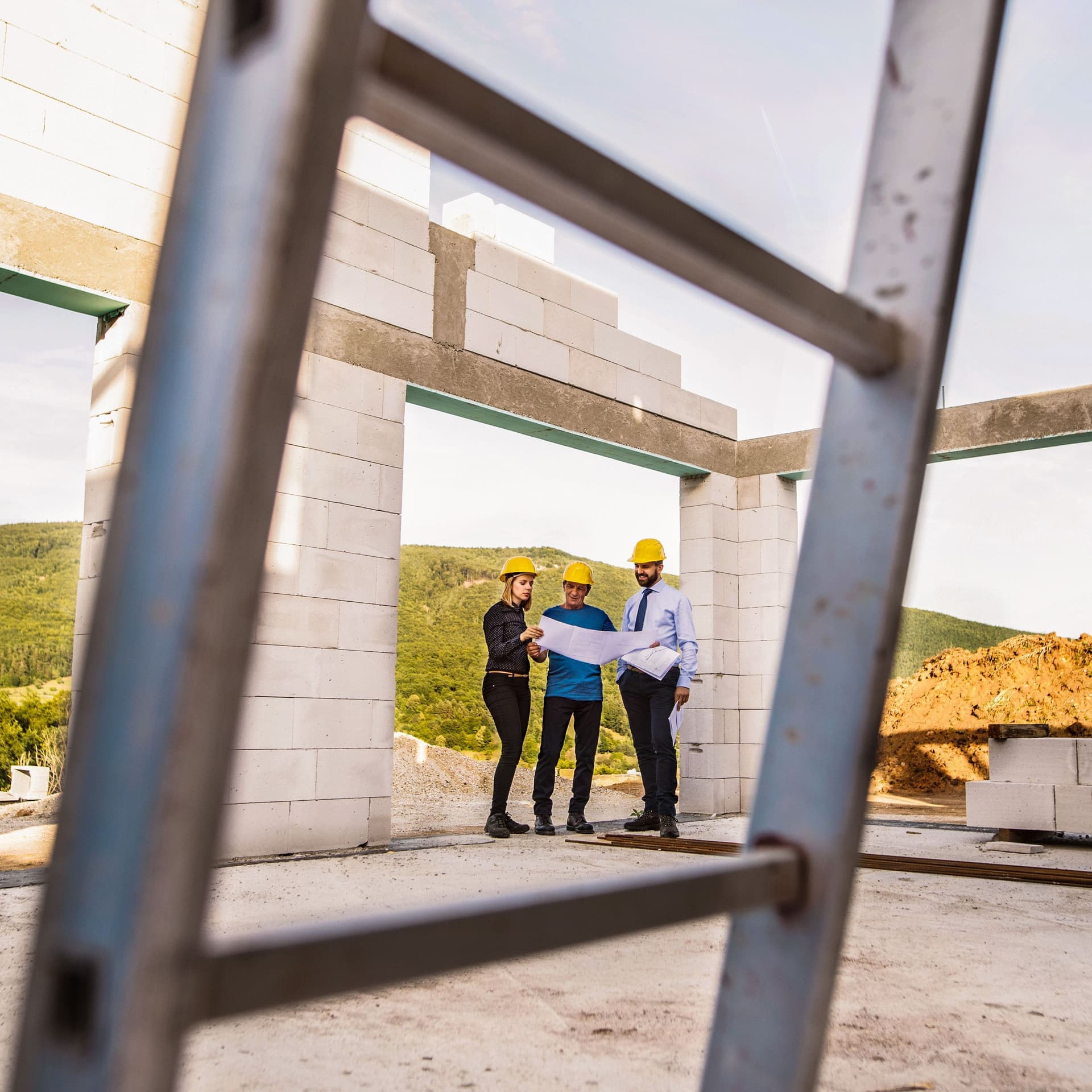 Drei Personen stehen mit gelben Helmen und Plänen auf Papier auf einer Baustelle eines Hauses. Im Vordergrund steht eine Metallleiter.