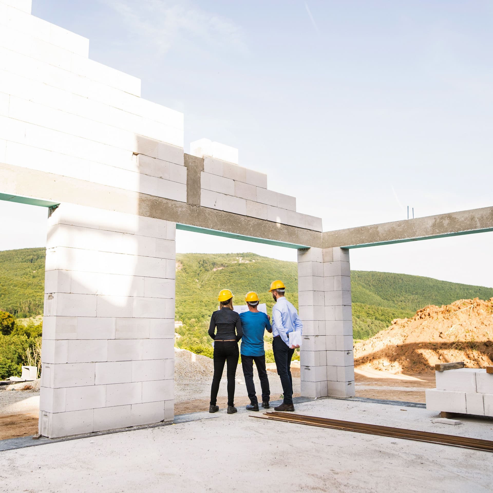 Drei Architekten und Arbeiter mit gelben Helmen und einem Plan in der Hand auf der Baustelle eines Rohbaus.