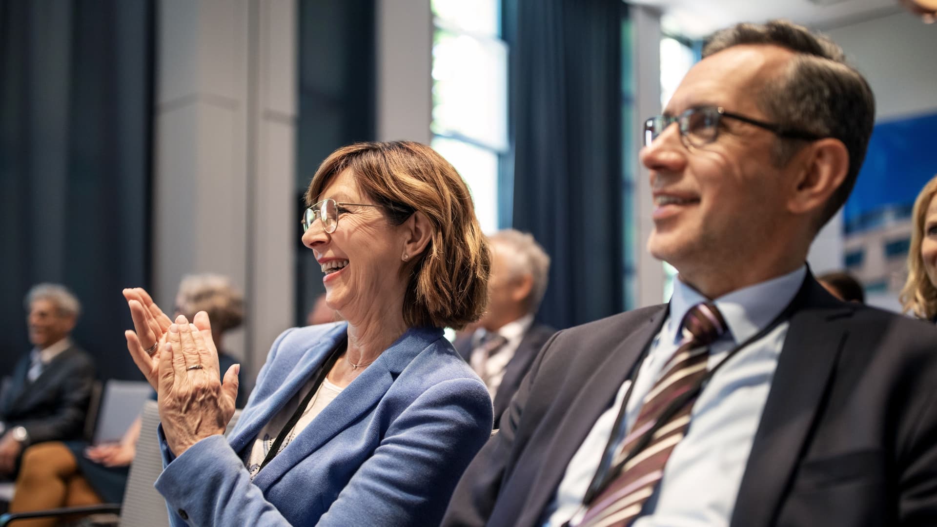 Lächelnde Frau applaudiert während eines Seminars. Sie sitzt zwischen anderen Teilnehmern. Alle tragen Business-Kleidung.