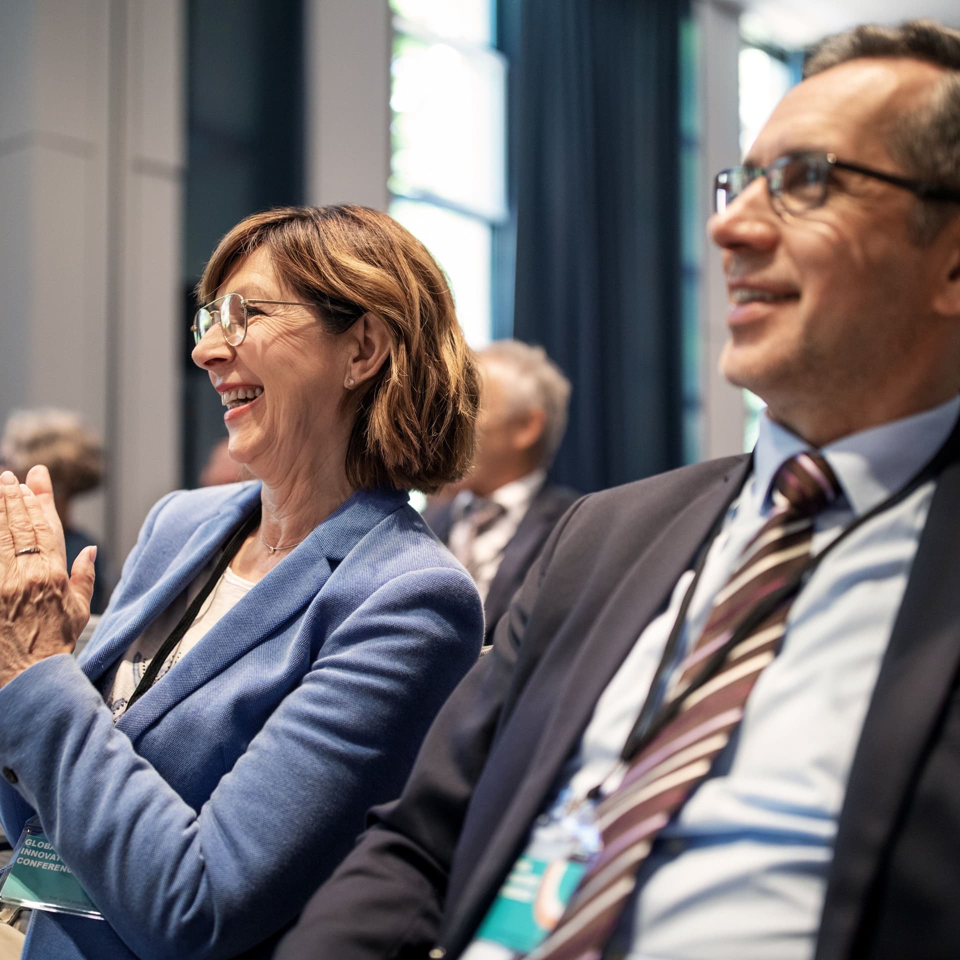 Lächelnde Frau applaudiert während eines Seminars. Sie sitzt zwischen anderen Teilnehmern. Alle tragen Business-Kleidung.