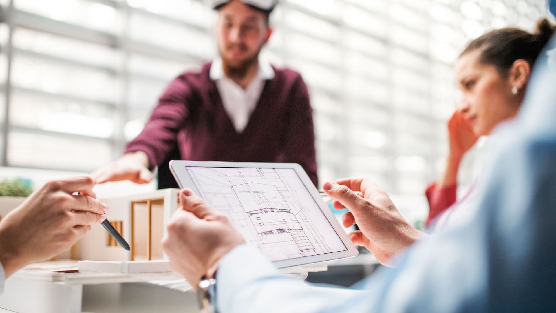 Eine Gruppe junger Architekten mit einem Tablet und einem Hausmodell steht in einem Büro und unterhält sich.