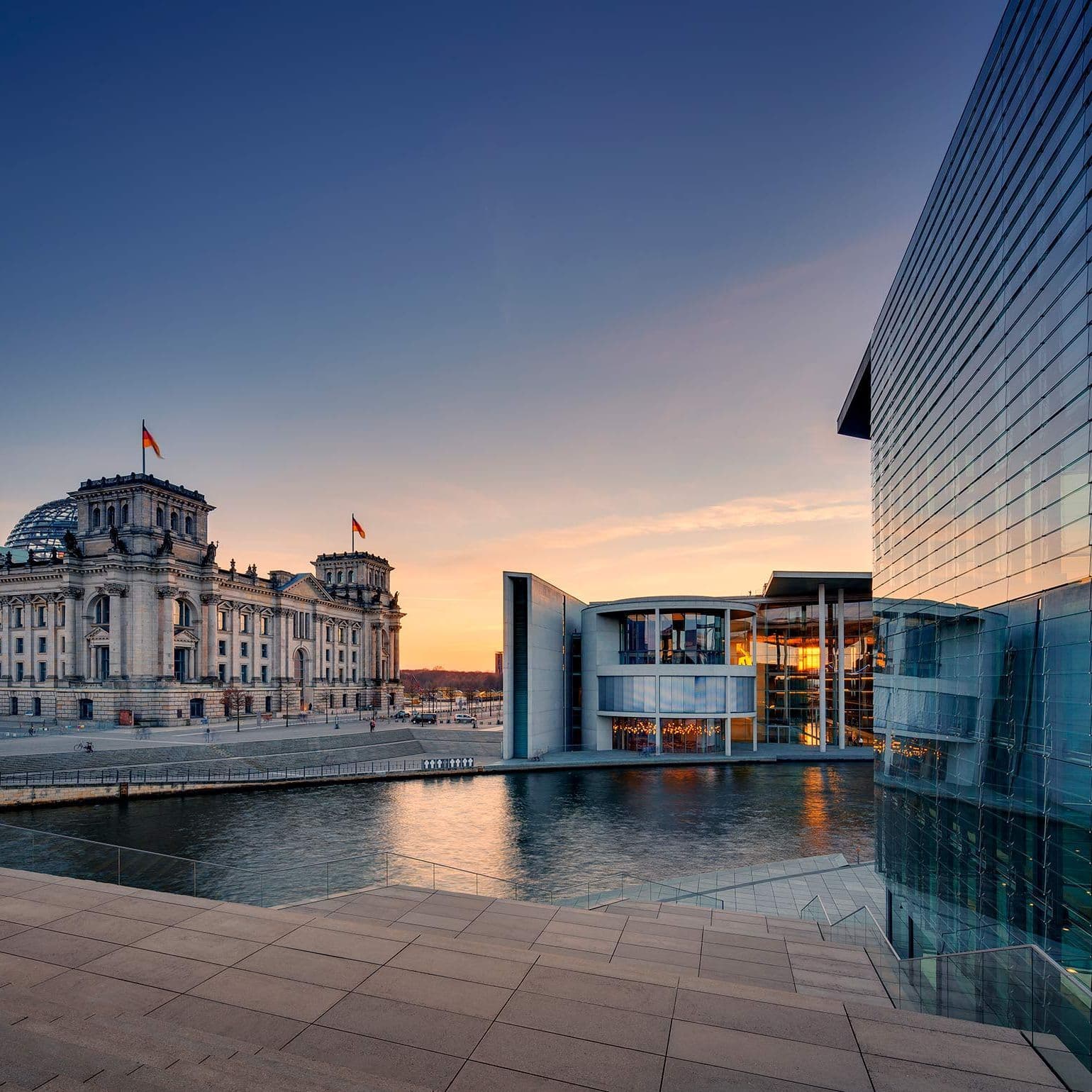 Blick auf den Reichstag mit dem Marie-Elisabeth-Lüders-Haus im Vordergrund. Es dämmert und die Regierungsbauten sind in ein blaues und rotes Licht getaucht.