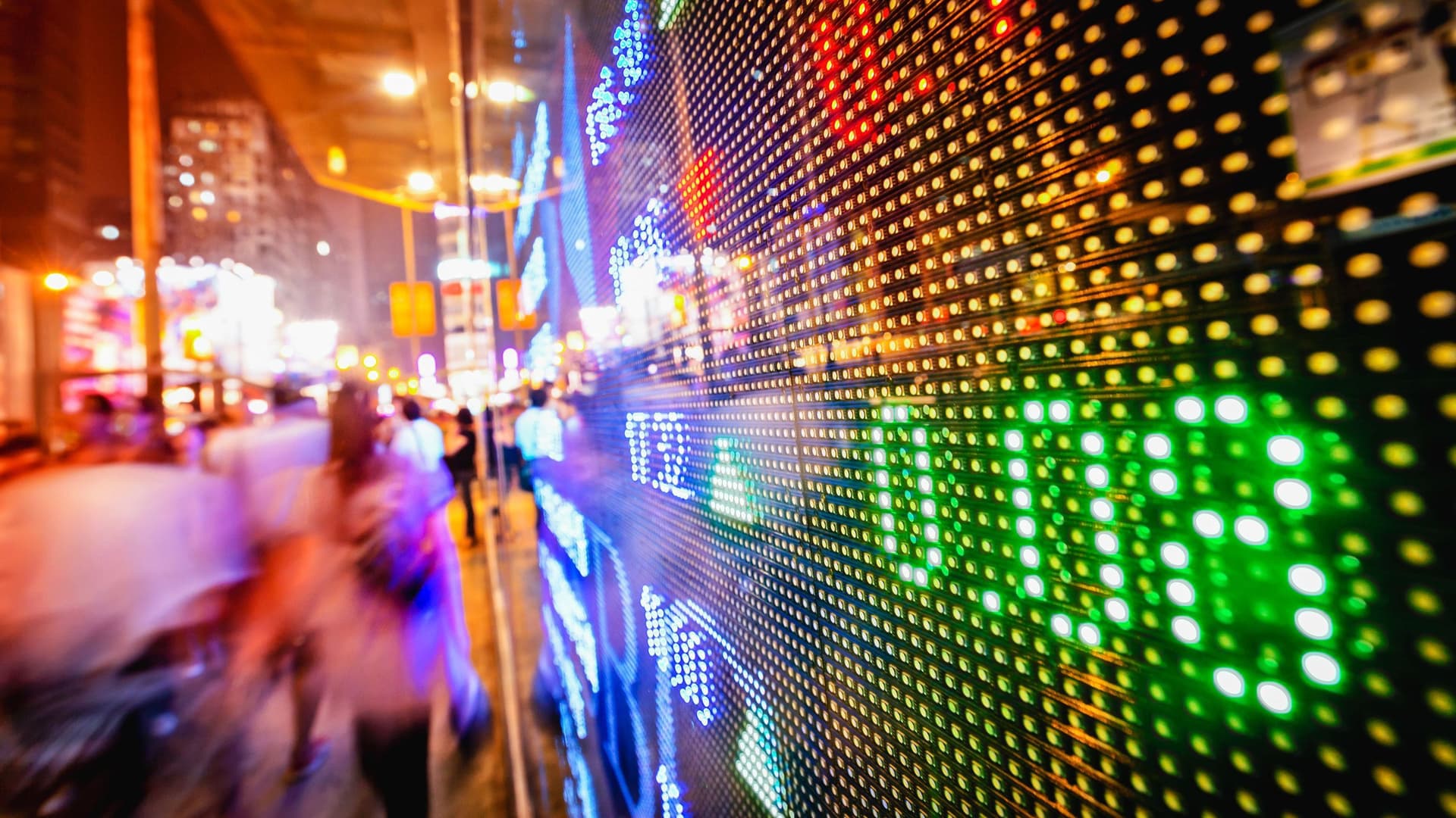 LED Anzeige von Börsenkursen auf einer großen Schaufensterfront. Unscharf laufen Menschen in einer Abendstimmung den Bürgersteig entlang.