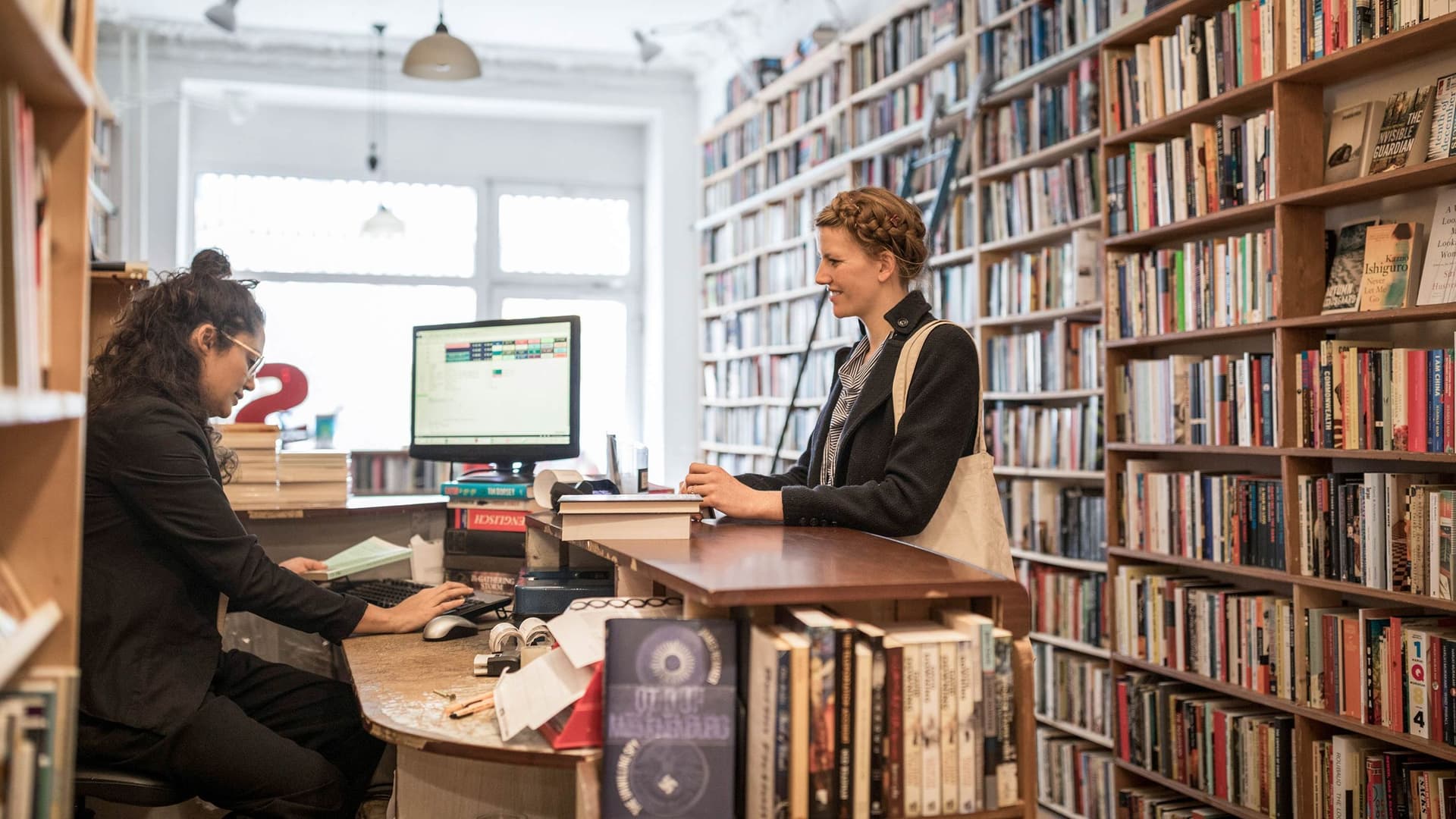 Eine junge Kundin steht am Tresen einer Buchhandlung, umrahmt von Bücherregalen. Sie schaut auf die sitzende Buchhändlerin, die etwas in einen Computer eingibt.