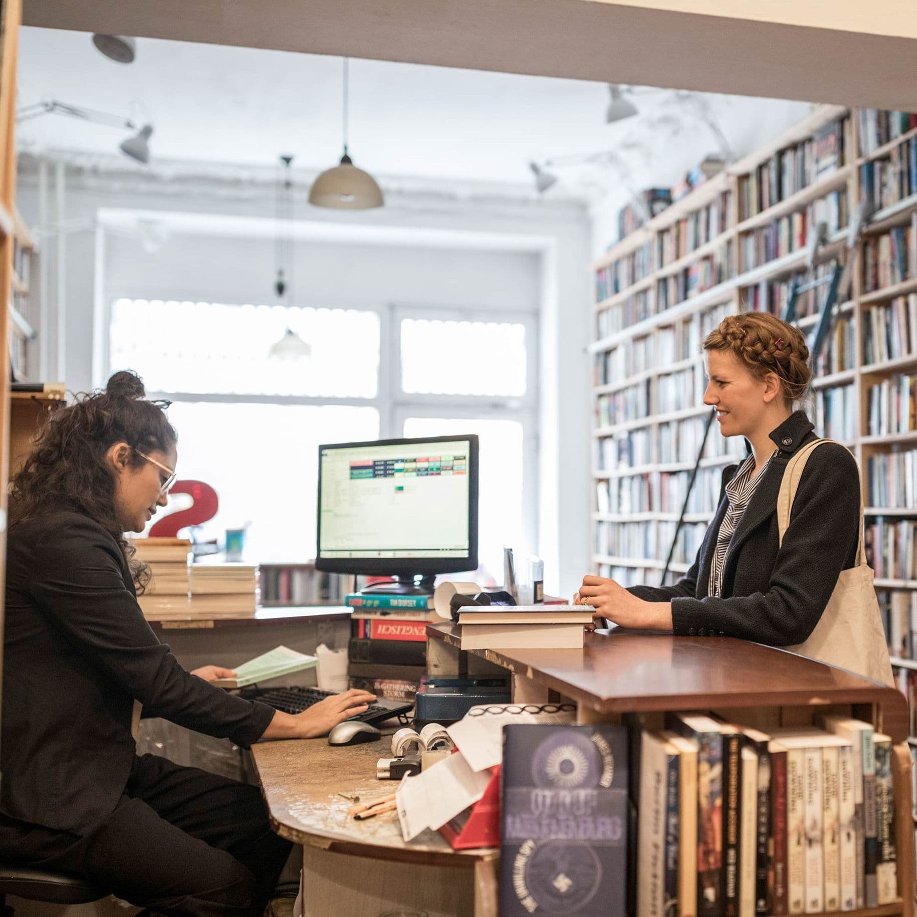 Eine junge Kundin steht am Tresen einer Buchhandlung, umrahmt von Bücherregalen. Sie schaut auf die sitzende Buchhändlerin, die etwas in einen Computer eingibt.