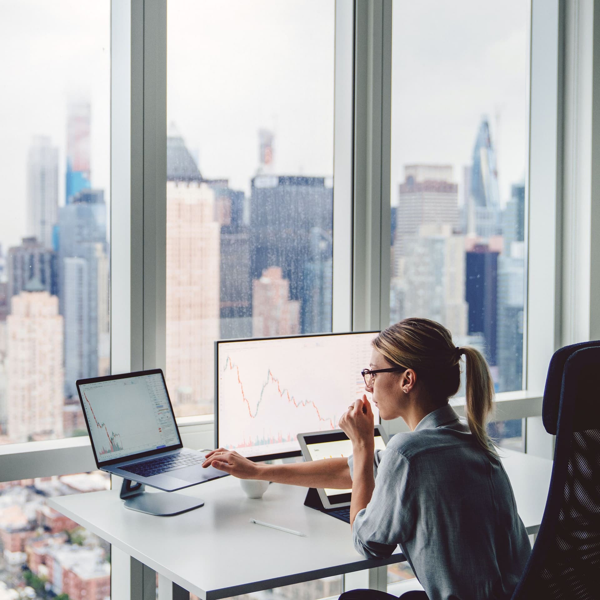 Konzentriert arbeitende Frau vor einem Panoramafenster sitzt an einem Schreibtisch mit zwei Bildschirmen. Im Hintergrund ist eine verschwommene Skyline zu erkennen.