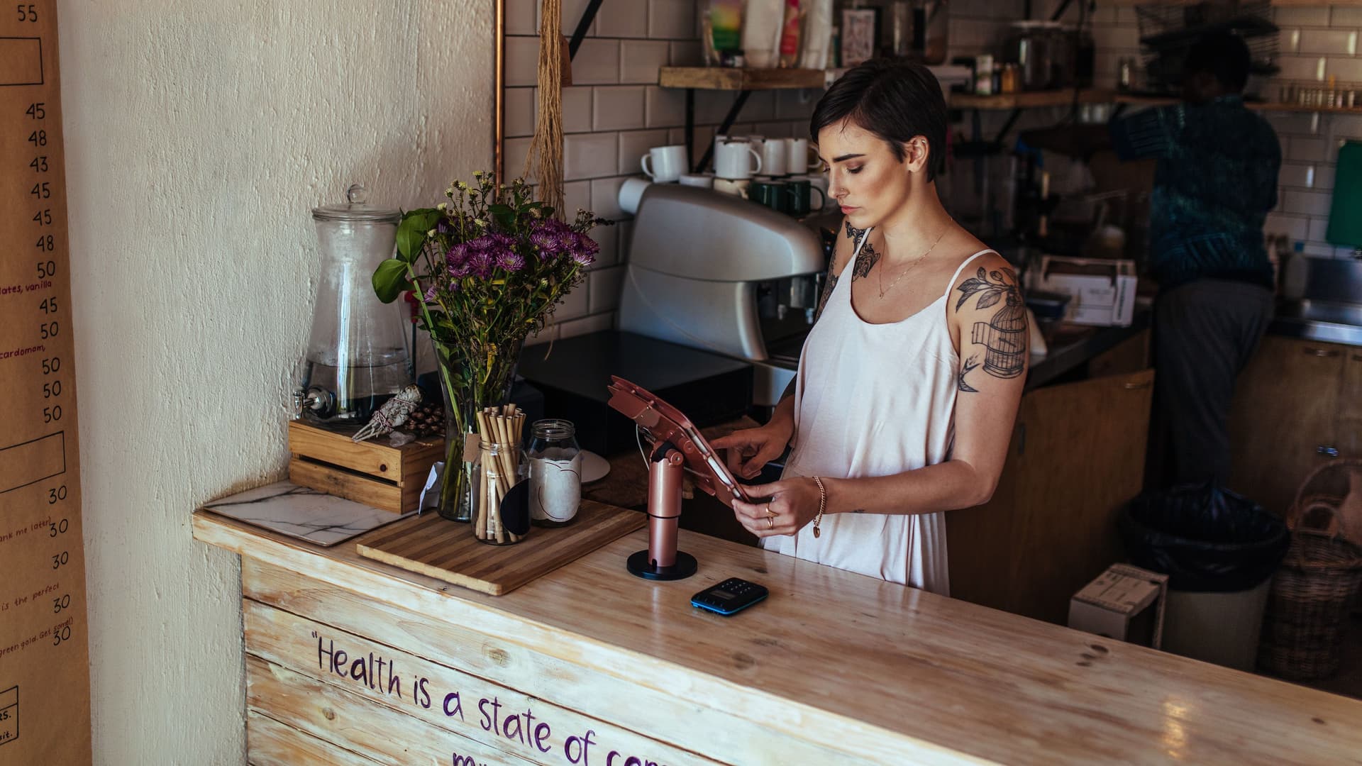 Junge Frau mit kurzen Haaren steht mit einem Tablet hinter dem Holz-Theresen in einem Café.