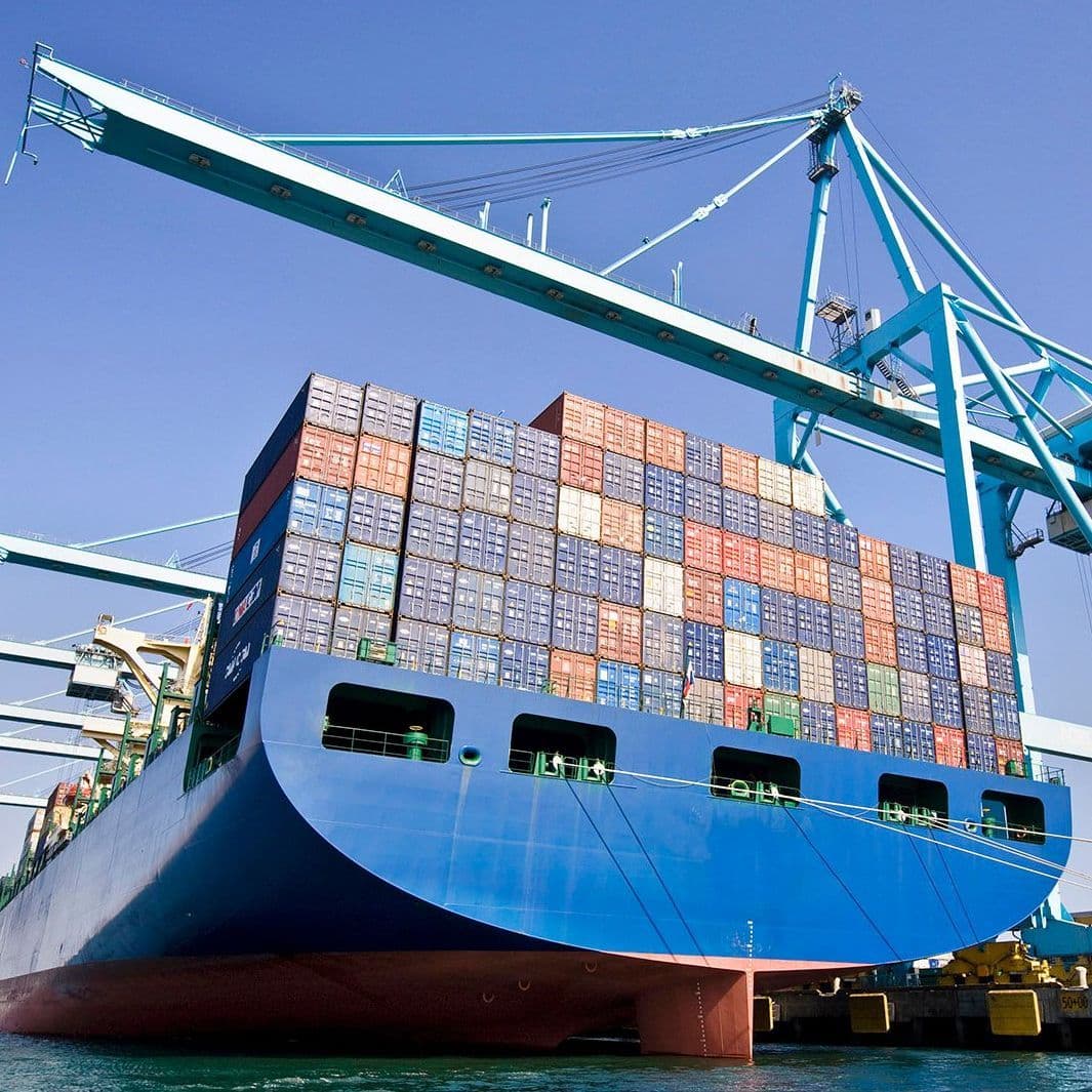 Ein großes Containerschiff liegt an einem Hafen. Über den bunten, gestapelten Containern ist ein Kran vor blauem Himmel zu sehen. 