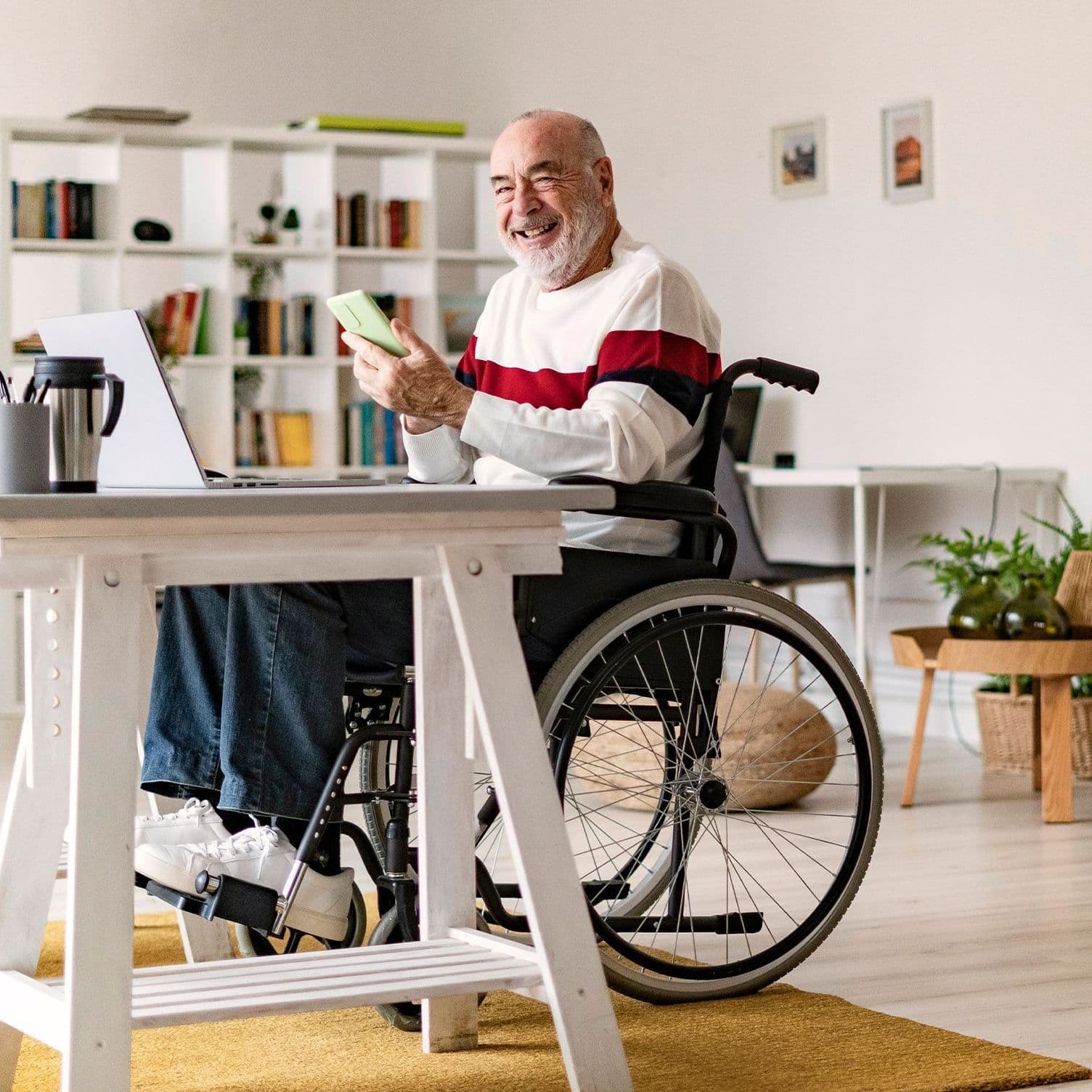 Älterer Mann im Rollstuhl sitzt mit seinem Smartphone an seinem Schreibtisch.
