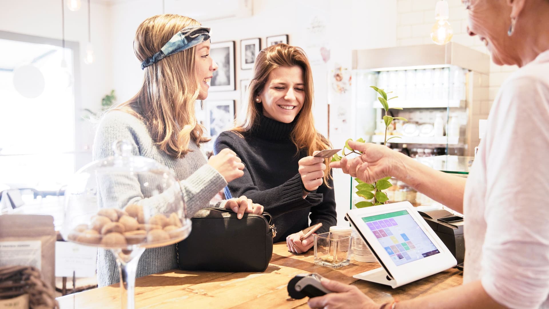 Zwei junge lachende Frauen stehen in einem Café an einem Holz-Tresen an der Kasse. Die eine Frau bezahlt bei einer Frau hinter der Kasse mit einer Karte.
