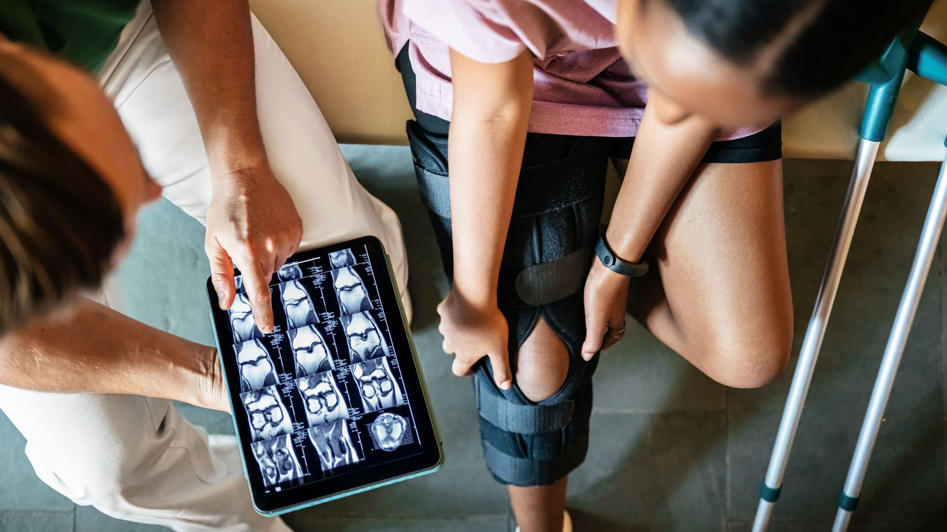 Bild von oben von einer Frau, die mit einer Orthese am Bein und Krücken neben sich auf einer Liege sitzt. Sie spricht mit einem Mann, der ein Tablet mit CT Bildern in der Hand hält.