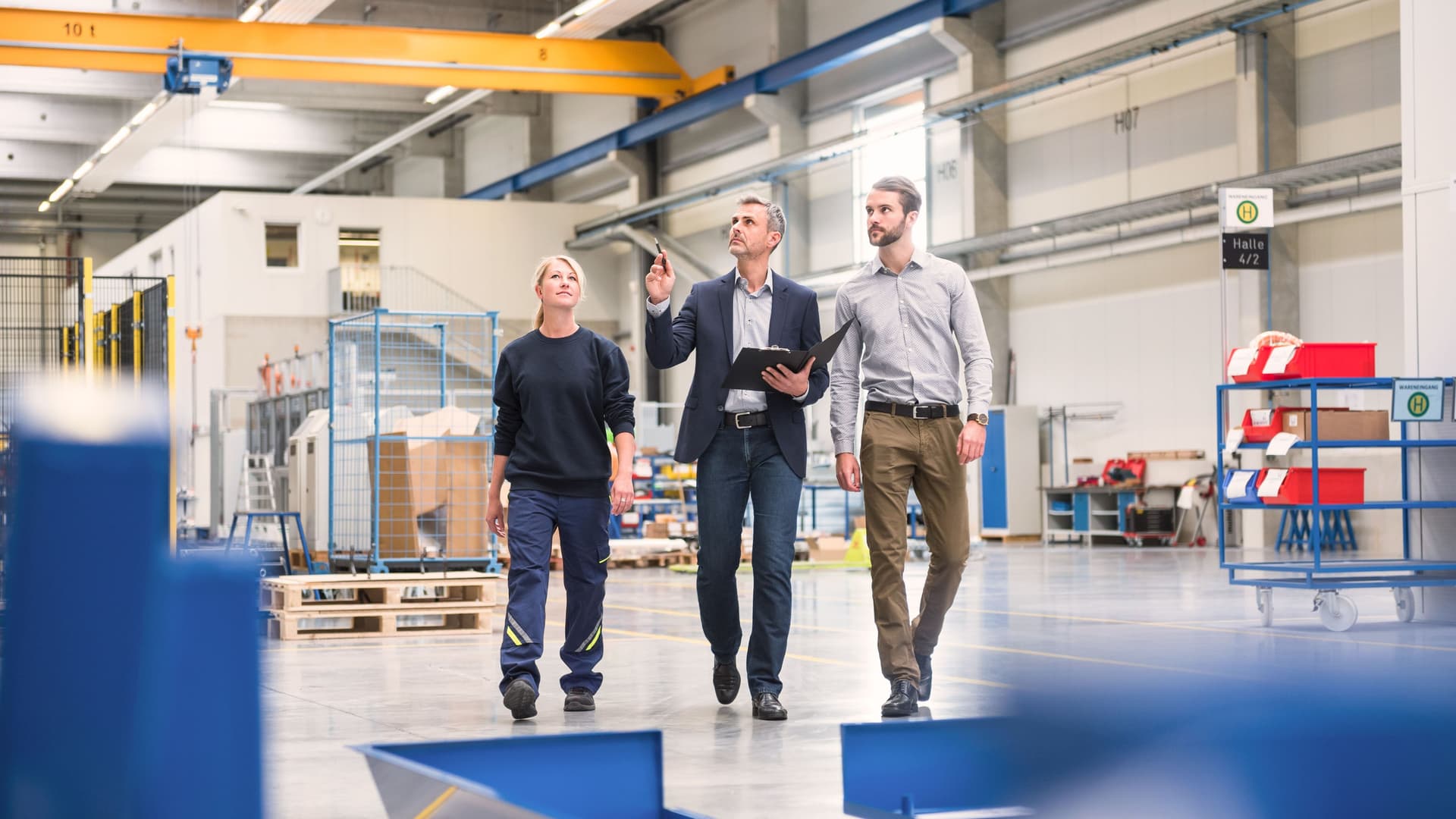 Zwei Männer und eine Frau schlendern durch eine Industriehalle und schauen nach oben. Der Mann in der Mitte hat eine Mappe und einen Stift in der anderen Hand.