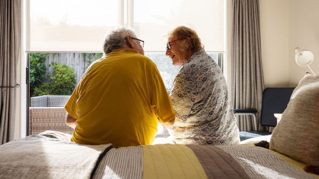 Ein älteres Paar sitzt auf dem Bett. Es scheint die Sonne. Sie lächeln sich an.