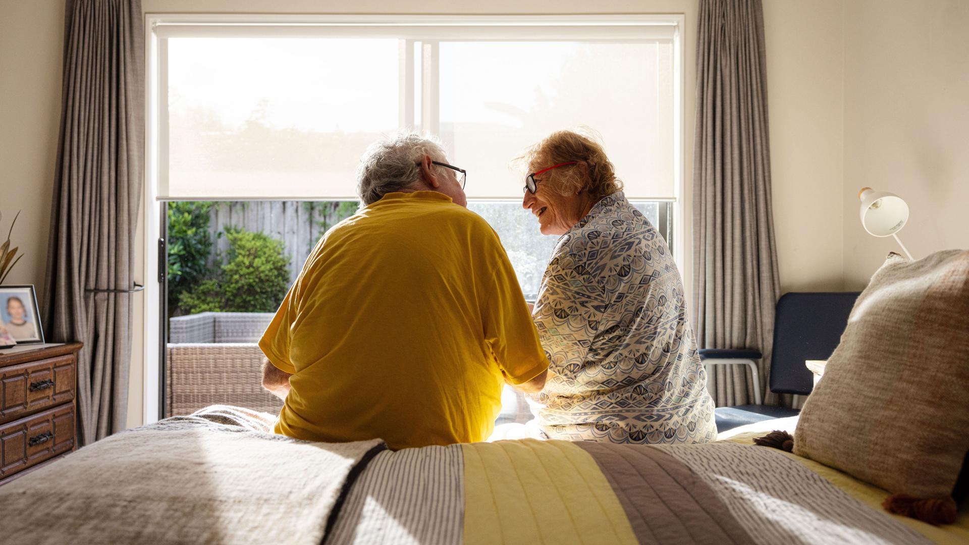 Ein älteres Paar sitzt auf dem Bett. Es scheint die Sonne. Sie lächeln sich an.