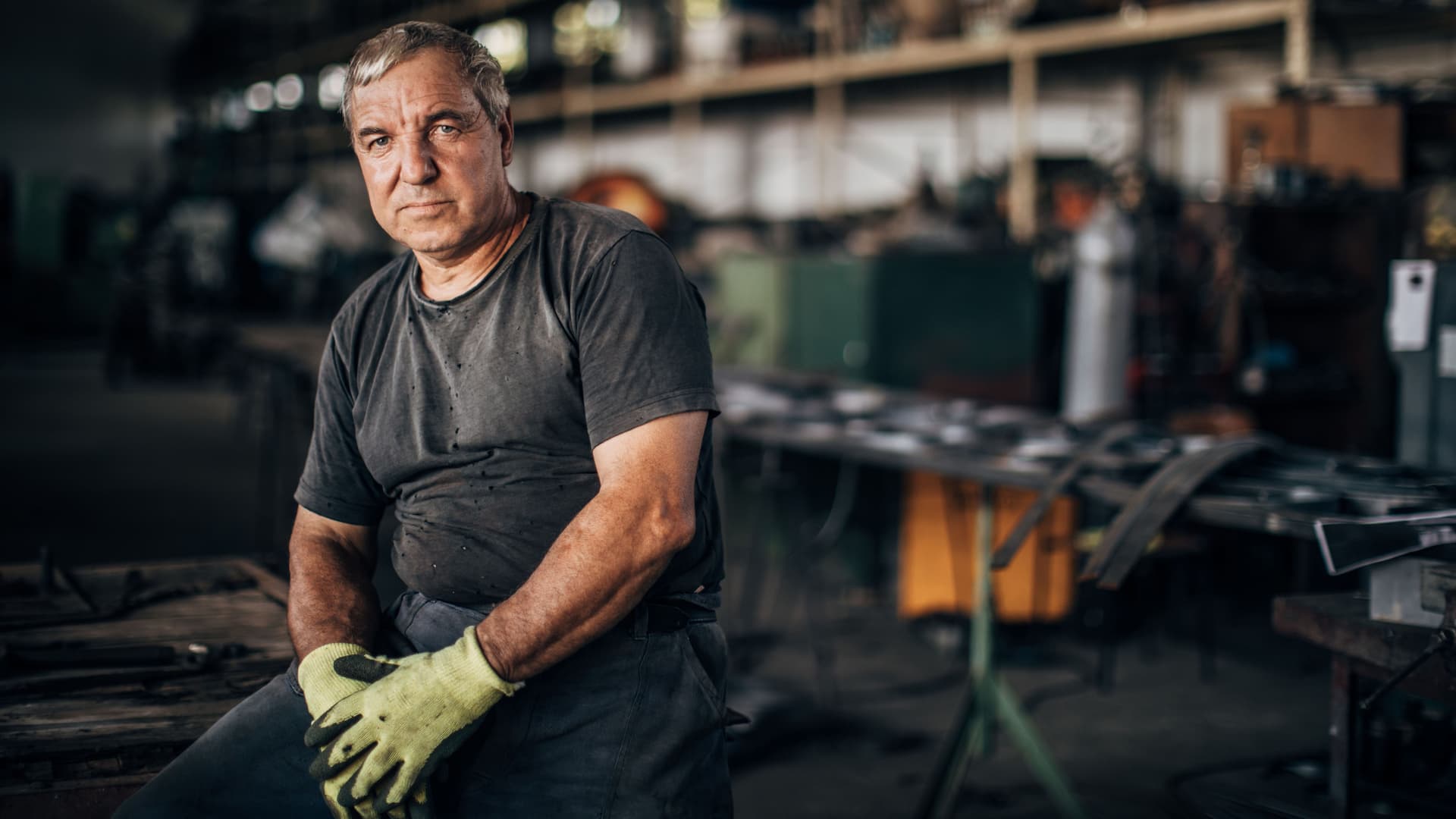 EIn Arbeiter macht eine Pause. Im Hintergrund ist die Werkstatt zu sehen. Er schaut genau in die Kamera.