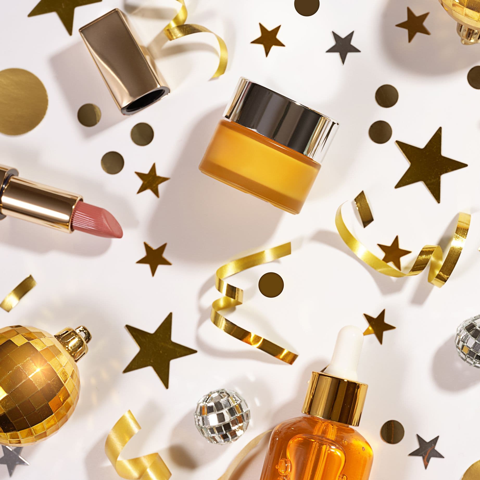 Goldene Kosmetikprodukte, festliche Deko wie Sterne, Konfetti und kleine Disco-Kugeln liegen kunstvoll arrangiert auf weißem Hintergrund und erzeugen eine elegante, feierliche Stimmung.