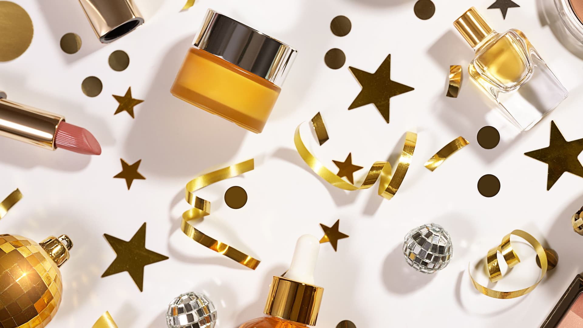 Goldene Kosmetikprodukte, festliche Deko wie Sterne, Konfetti und kleine Disco-Kugeln liegen kunstvoll arrangiert auf weißem Hintergrund und erzeugen eine elegante, feierliche Stimmung.