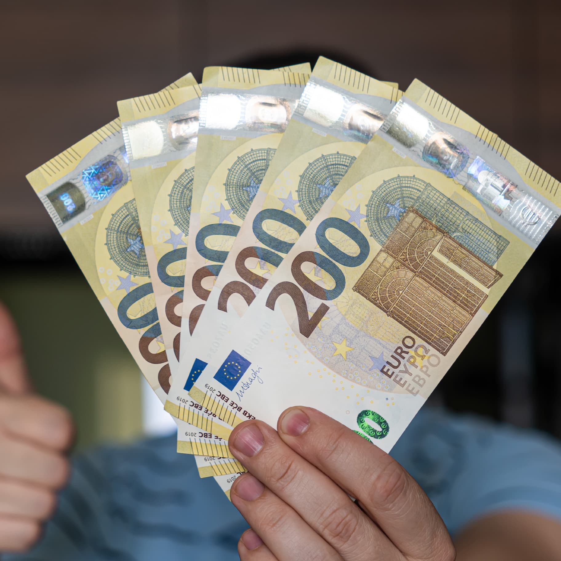 Eine Nahaufnahme zeigt eine Hand, die fünf 200-Euro-Banknoten fächerförmig hält. Die Banknoten sind goldfarben und zeigen typische Euro-Sicherheitsmerkmale. Im Hintergrund ist leicht unscharf ein Teil eines Daumens zu erkennen, der nach oben zeigt.