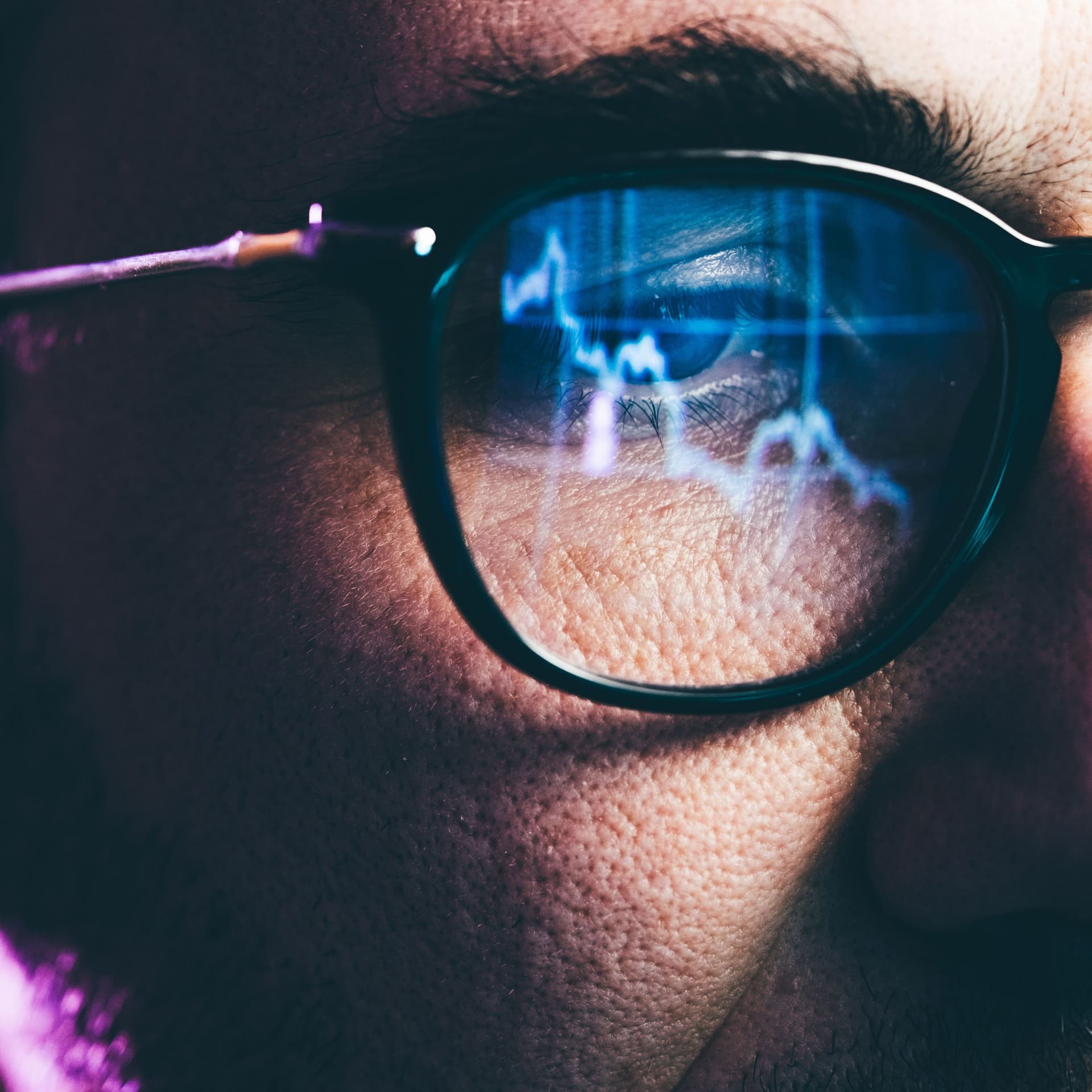 Nahaufnahme eines Mannes mit Brille. In seiner Brille spiegeln sich Aktienkurse.