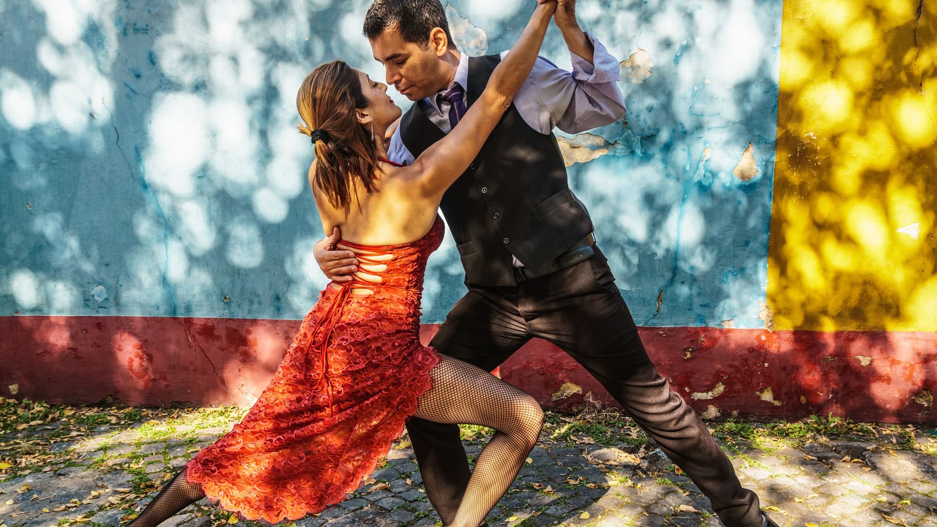 Ein Paar tanzt leidenschaftlich Tango auf einer Kopfsteinpflasterstraße; die Frau im roten Kleid und der Mann im dunklen Anzug halten eine dynamische Pose vor einer weißen Wand.