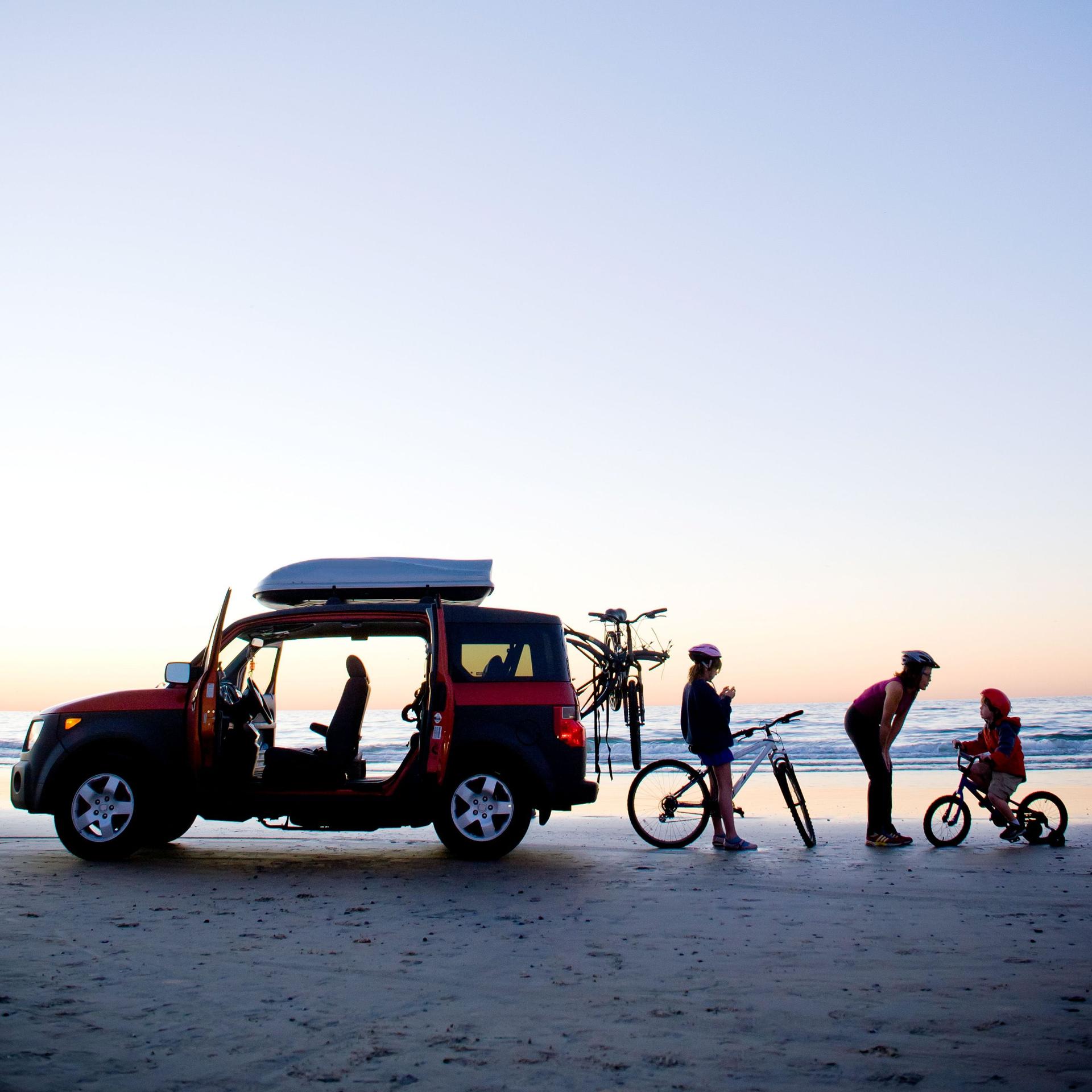 Eine Familie steht mit einem Auto und ihren Fahrrädern am Meer. Im Hintergrund ist die Sonne gerade untergegangen.