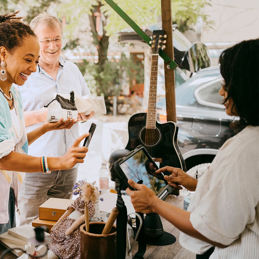 Eine Frau bezahlt auf einem Flohmarkt mit dem Smartphone, während eine Verkäuferin ein Kartenlesegerät hält. Im Hintergrund stehen ein Mann, eine Gitarre und verschiedene Secondhand-Artikel.