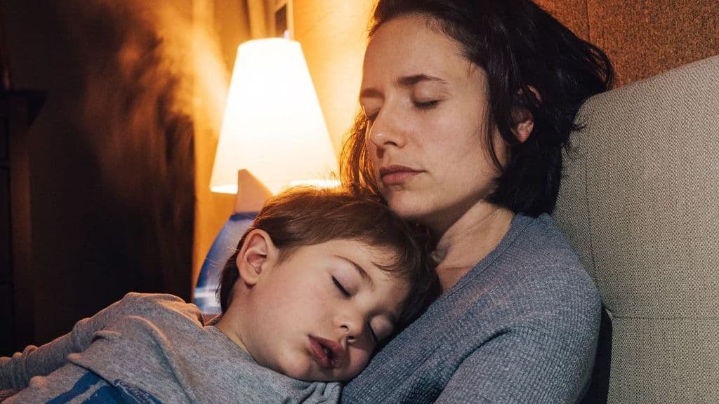 Eine Frau mit geschlossenen Augen sitzt auf dem Bett. Sie hält ihr krankes Kind im Arm.