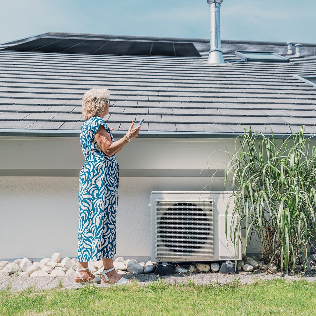 Eine Frau in blau-weißem Kleid steht vor einem Haus mit Solardach, blickt nach oben und hält ein Smartphone in der Hand.